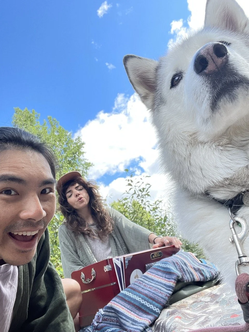 Two people and a large white dog outdoors on a sunny day with blue sky and clouds in the background.