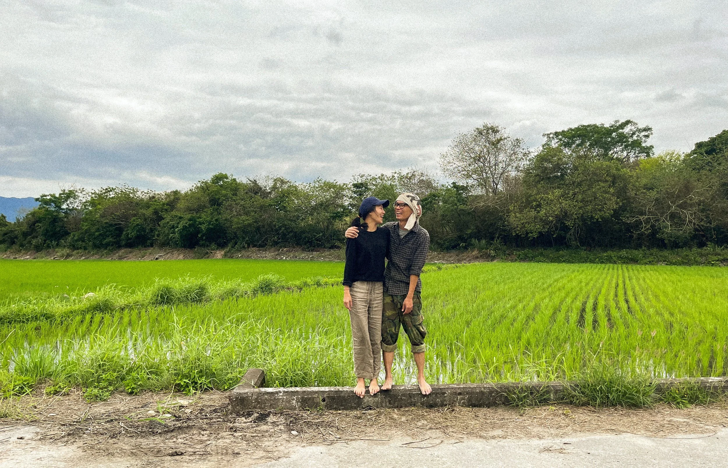 Two people standing barefoot beside a lush green rice field with trees in the background, under a cloudy sky.