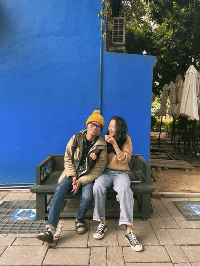 Two people sitting on a bench, smiling and interacting, with a bright blue wall in the background. One person is wearing a yellow knit hat and glasses, while the other has wavy hair and is wearing casual clothing.