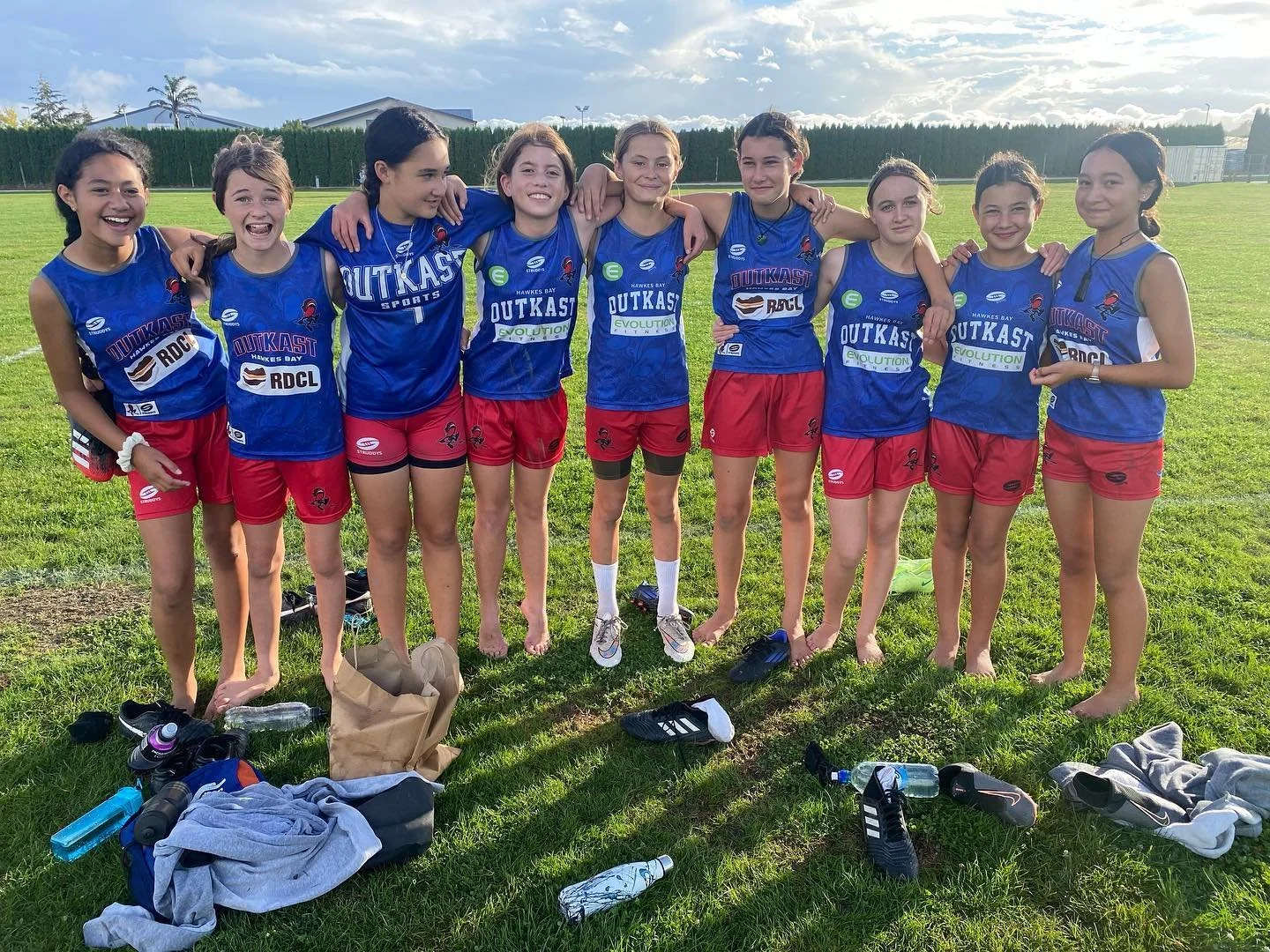 A group of nine young female athletes in blue and red sports uniforms standing arm-in-arm on a grassy field, smiling after a sports event on a sunny day.