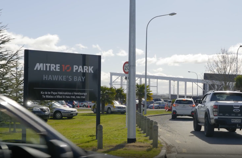 Sign at Mitre 10 Park in Hawke's Bay indicating it is a parking area, with cars parked and driving nearby, and a partly cloudy sky above.