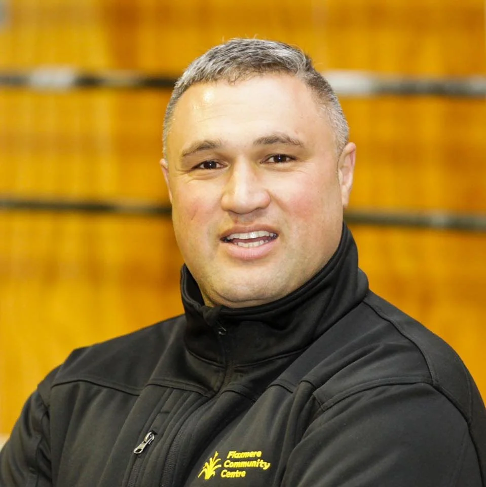 A man with short gray hair and a black jacket with yellow embroidery that reads 'Fluxmore Community Centre' with a yellow tree logo, standing indoors in front of a wooden wall.