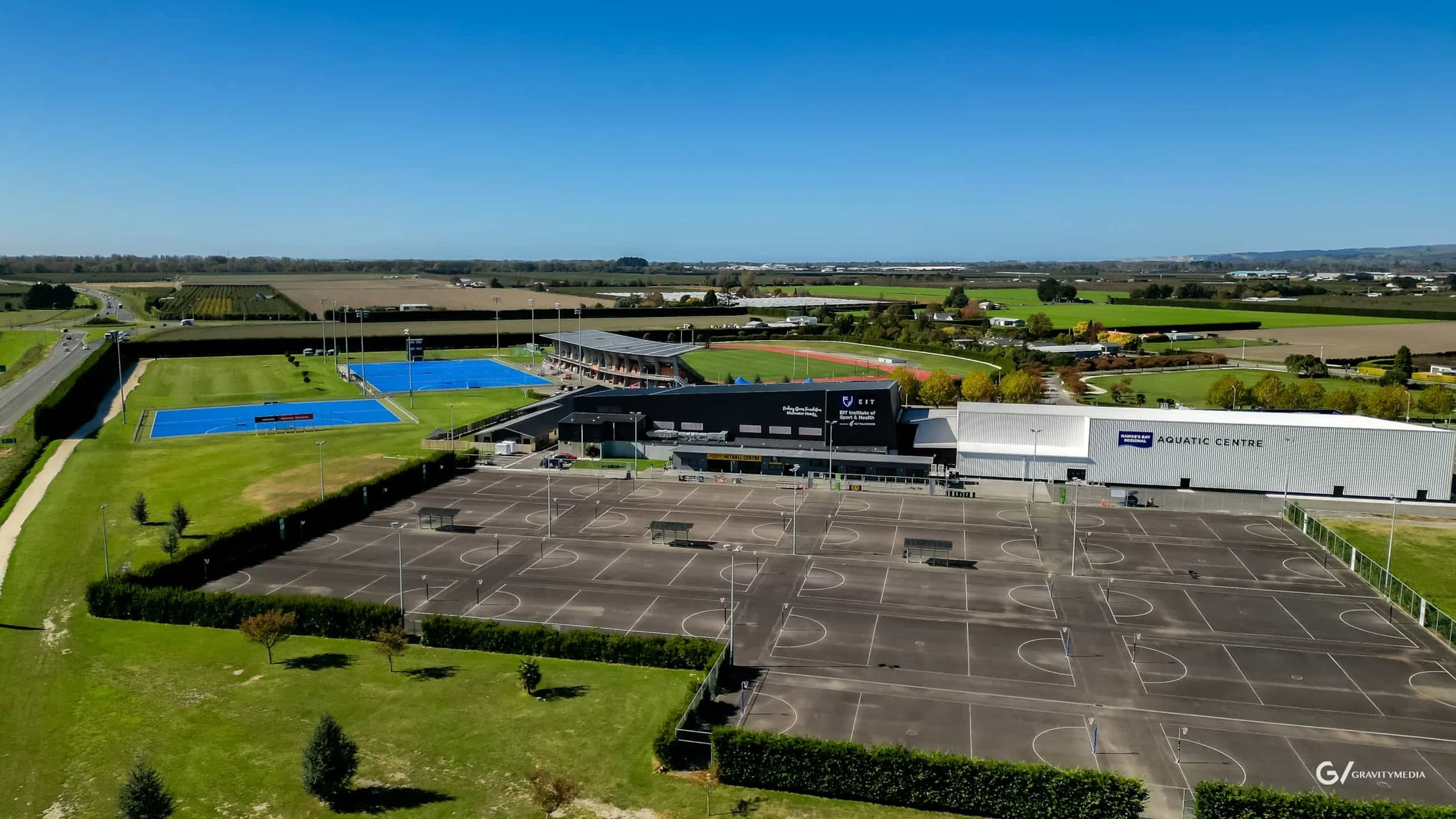 An aerial view of a sports complex showing tennis courts, a swimming pool, a building labeled 'Aquatic Centre,' and a parking lot with no cars, surrounded by green fields and open countryside.