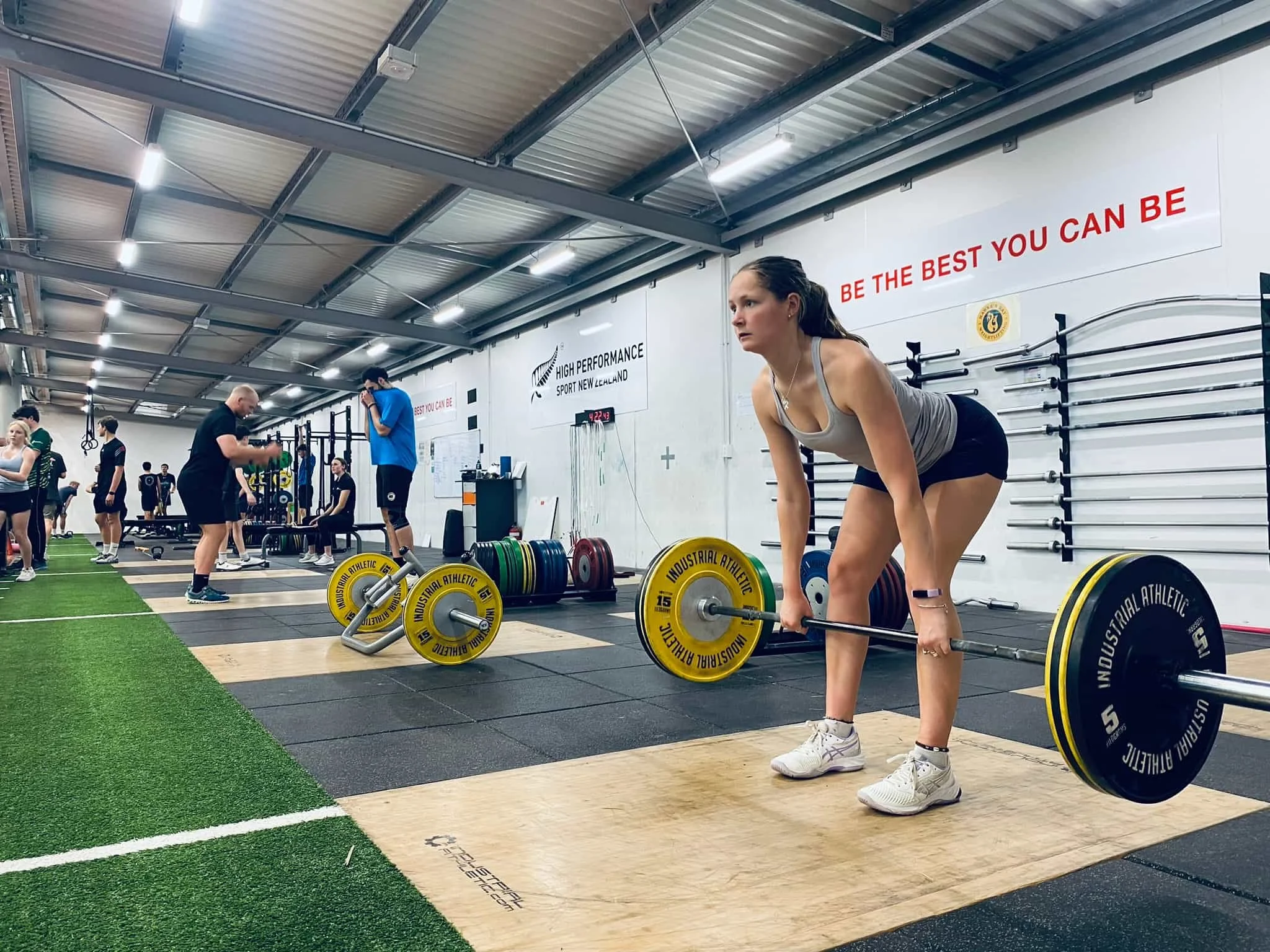 A woman lifting a barbell during a workout in a gym, with other people exercising in the background.