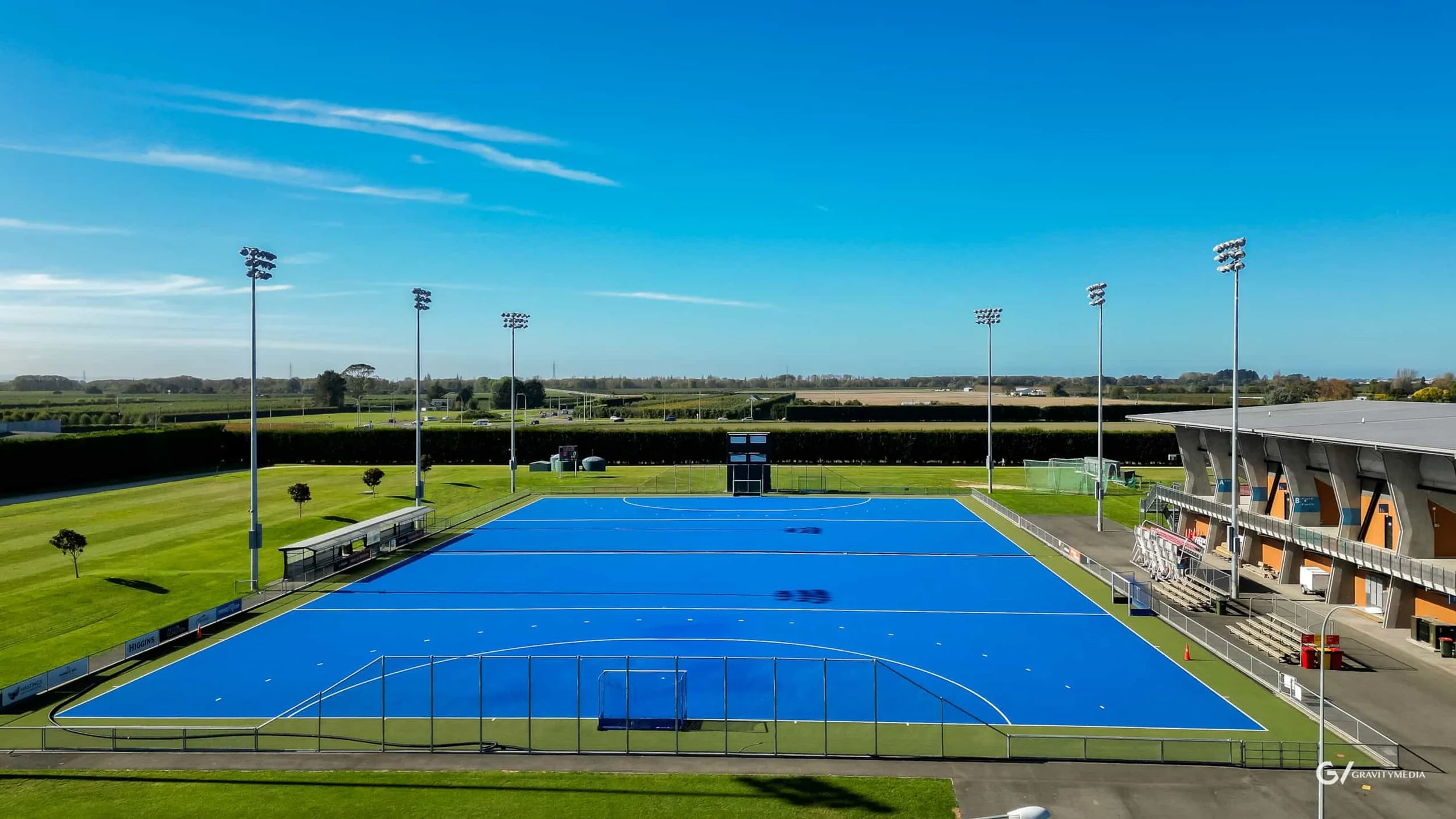 An outdoor sports field with a blue hockey pitch, surrounding green grass, tall light poles, a covered bench area, and a modern stands building under a clear blue sky.