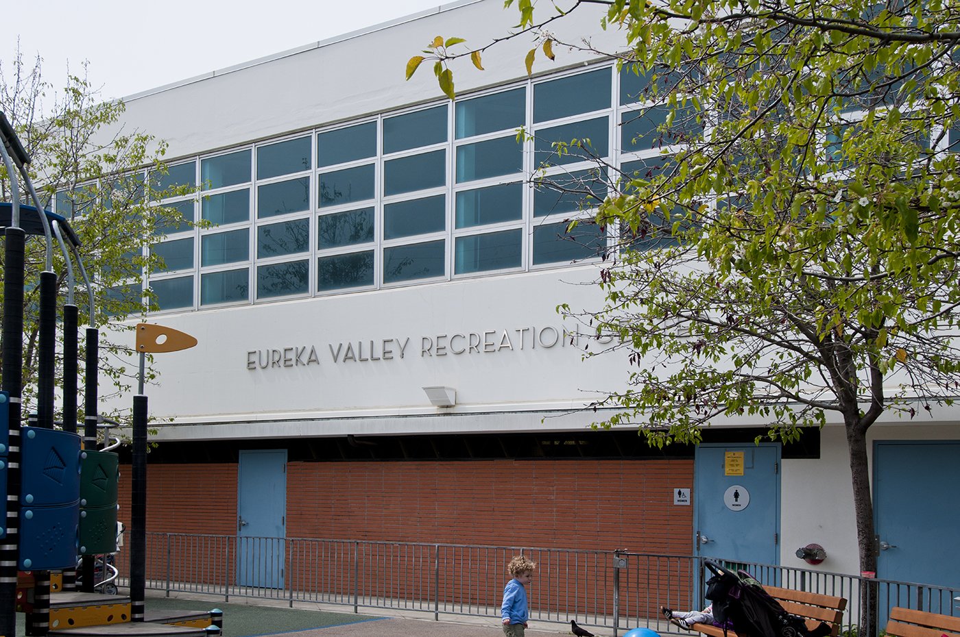 Eureka Valley Recreation Center building exterior with large windows, playground equipment, and a child playing in the foreground.