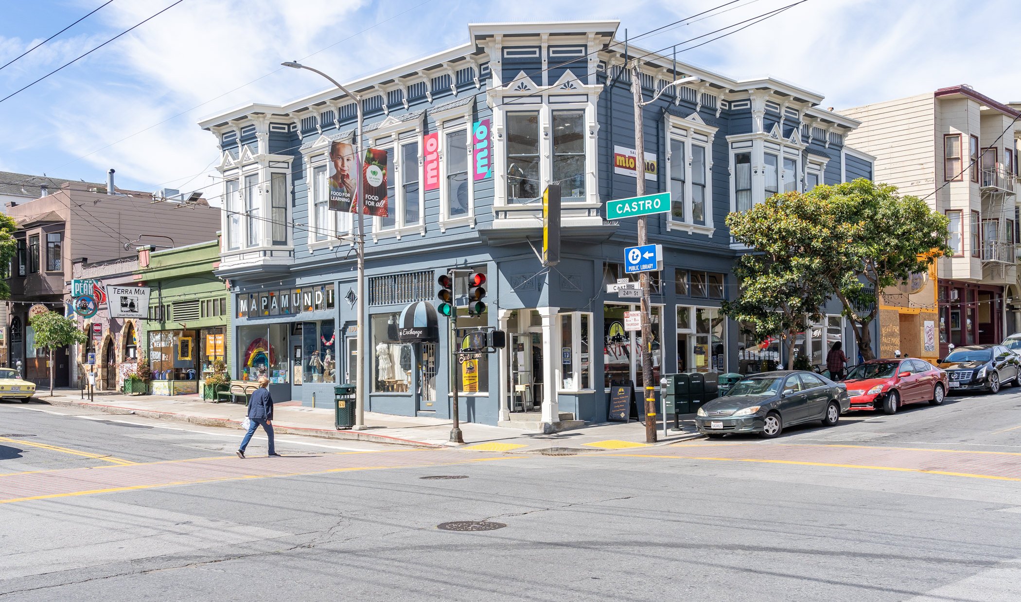 Street corner in a city with a Victorian-style building, featuring shops like "MAPAMUNDI" and "Terra Mia," with cars parked and a person crossing the street.