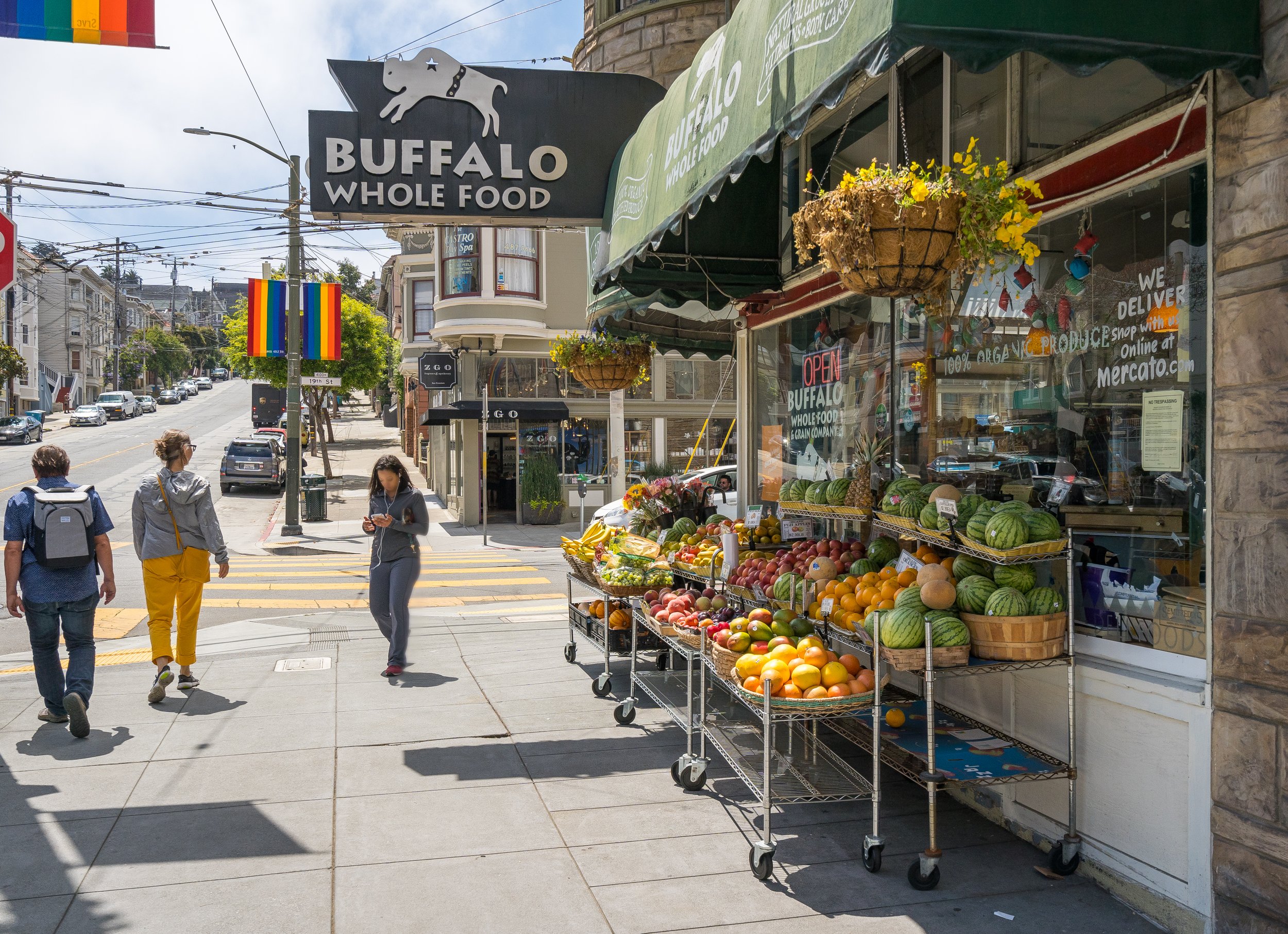 Street view of Buffalo Whole Food store with fruit stand, rainbow flags, and people walking.