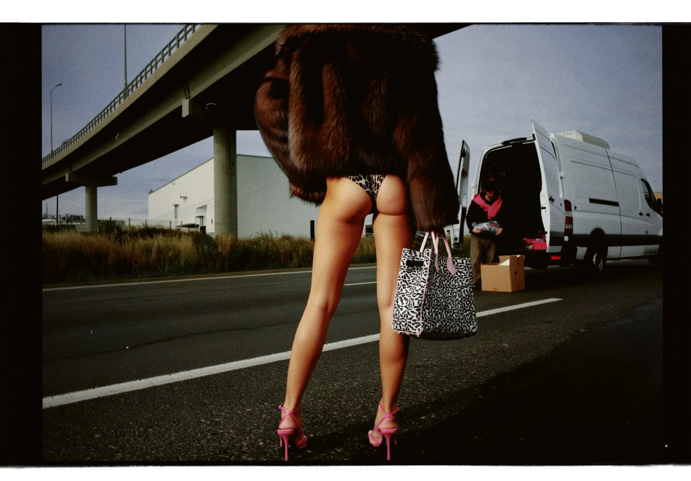 A woman in high heels and revealing clothing stands on the side of a highway, with a large fur coat and animal print bags nearby, as a person sorts items from a van in the background.