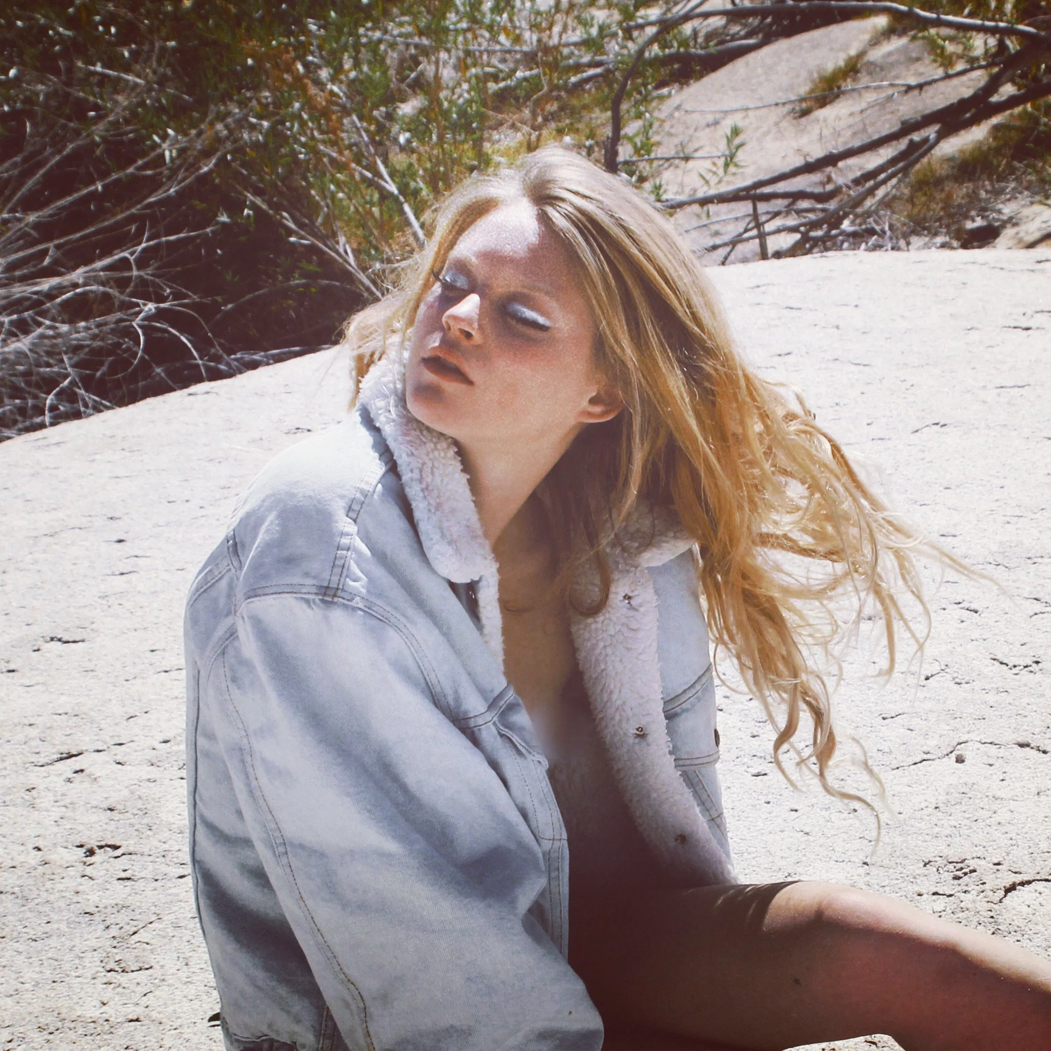 A young woman with blonde curly hair sitting outdoors on sand, wearing a denim jacket with a shearling collar, looking to her side with a serious expression.