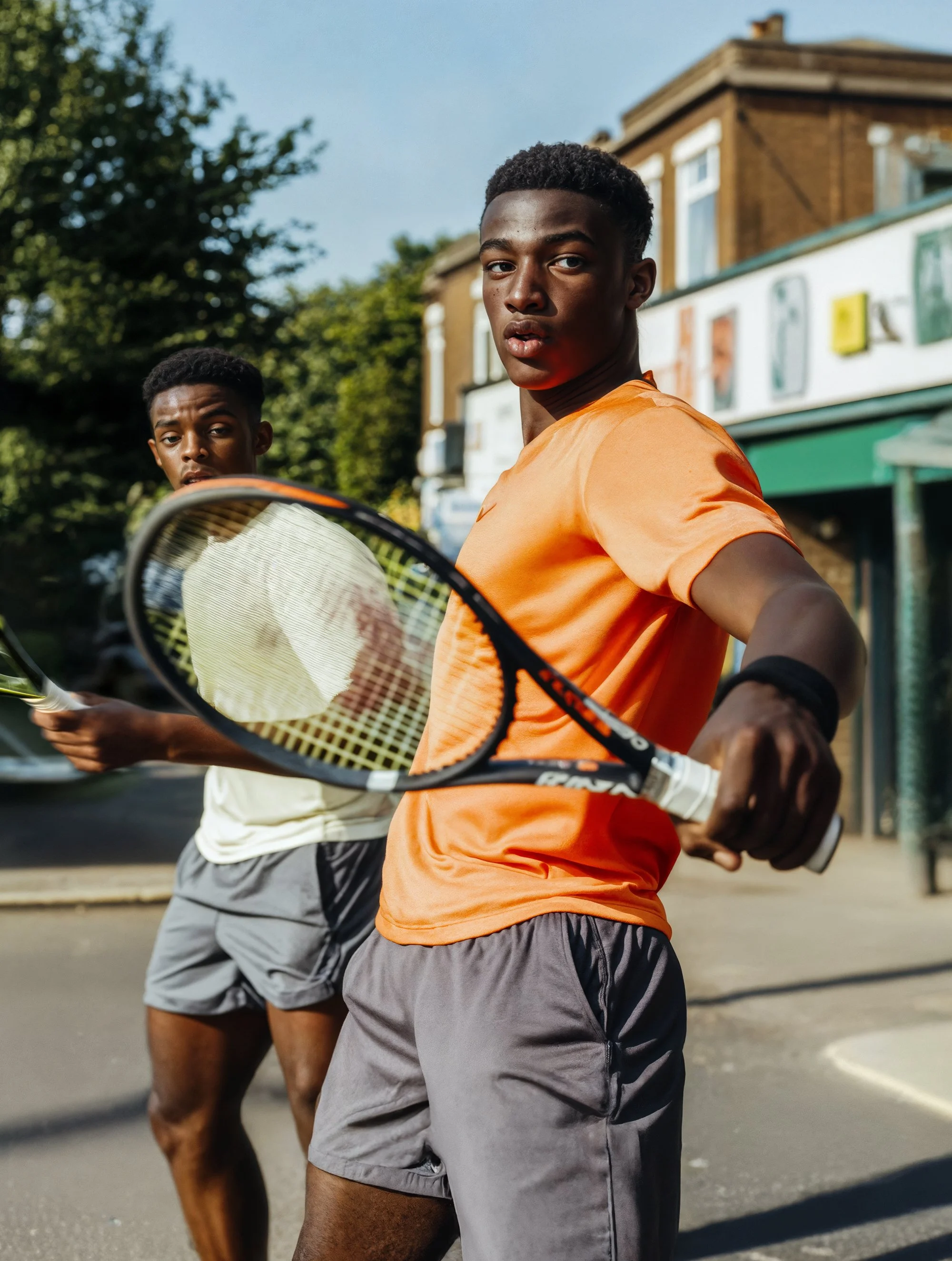 Two young men playing tennis outdoors on a city street, holding tennis rackets, with buildings and trees in the background.