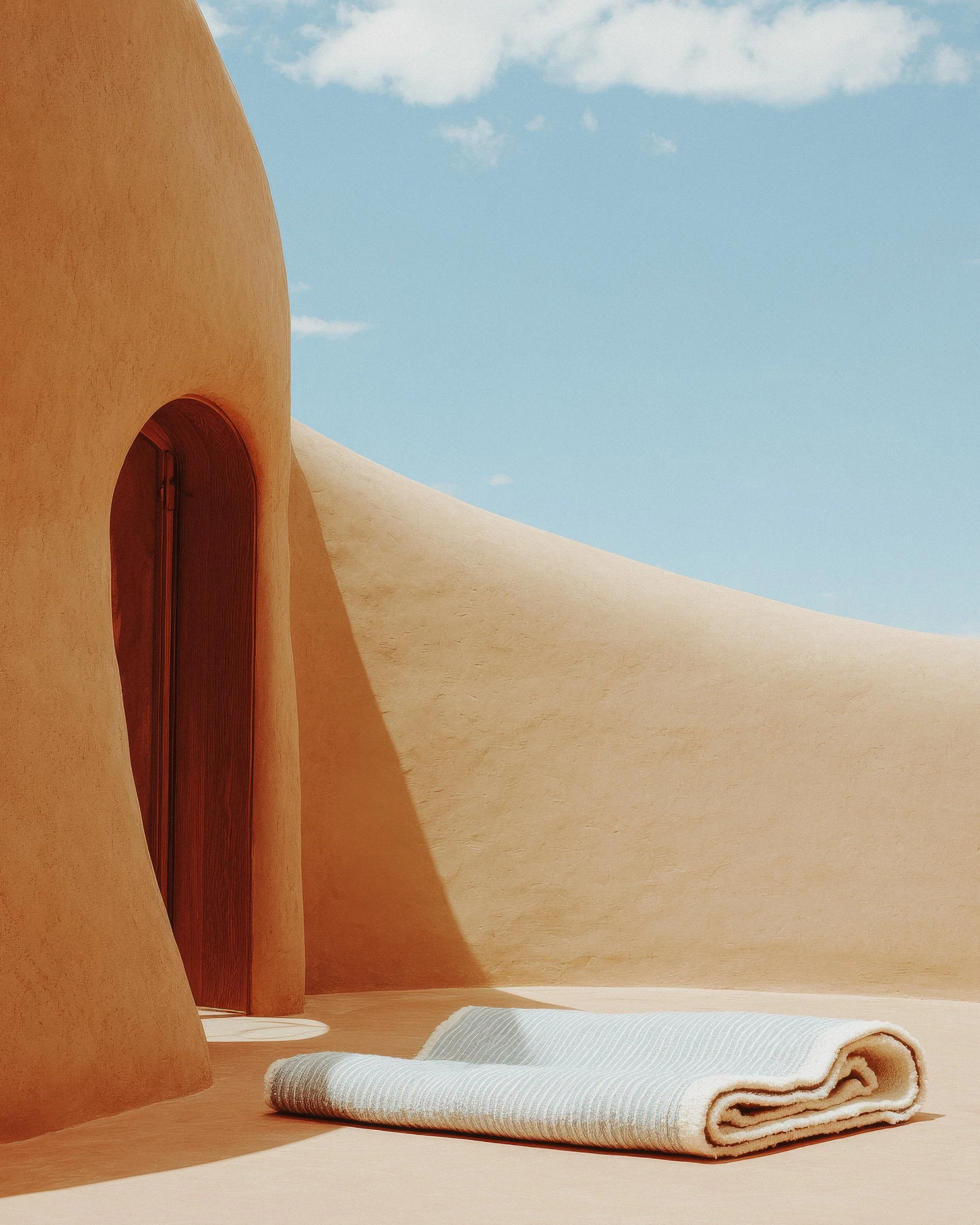Minimalist desert scene with smooth, curved beige structures, a rolled white towel, and a clear blue sky with scattered clouds.