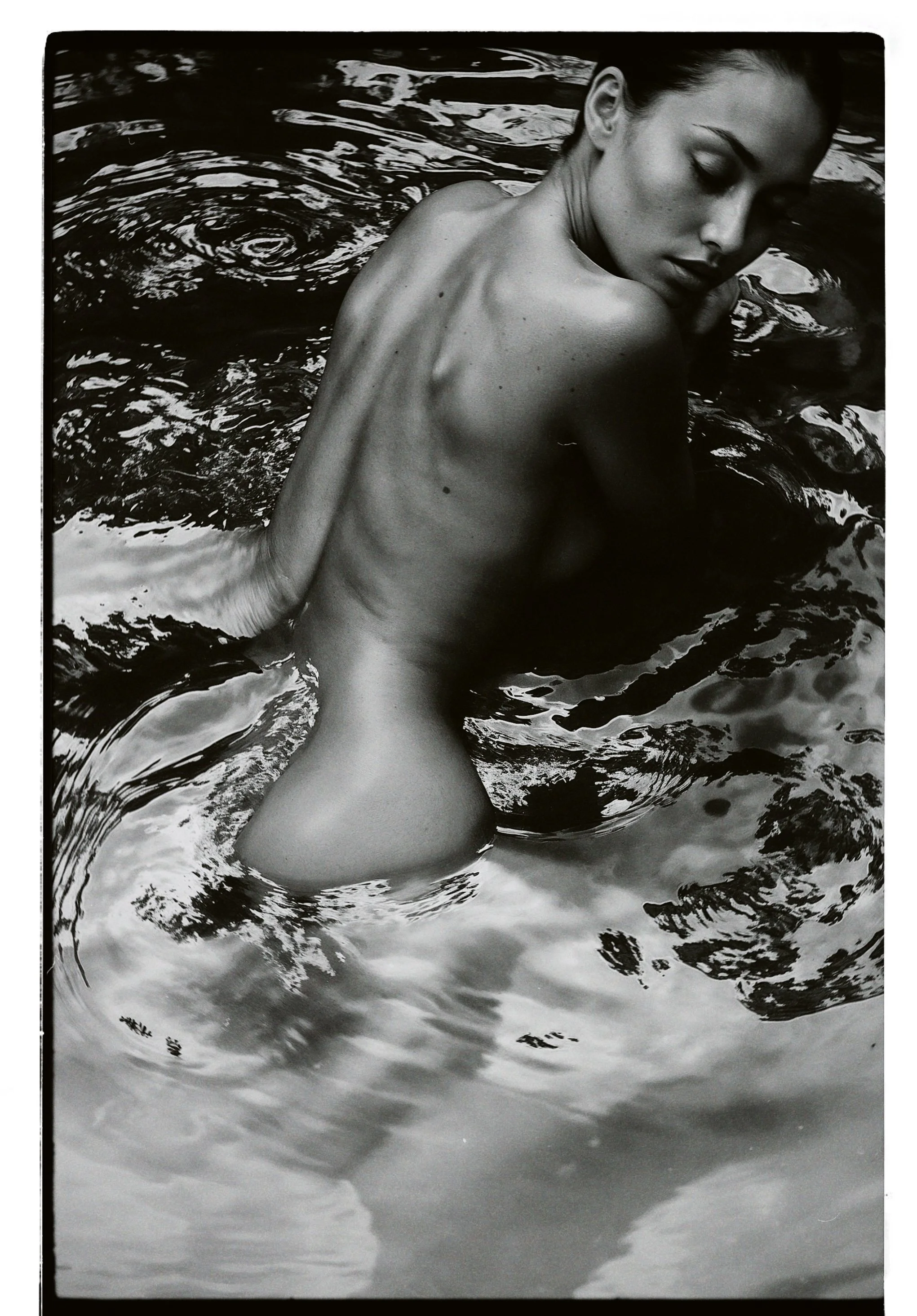Black and white photo of a nude woman with her eyes closed, partially submerged in water, with her back and shoulder exposed.