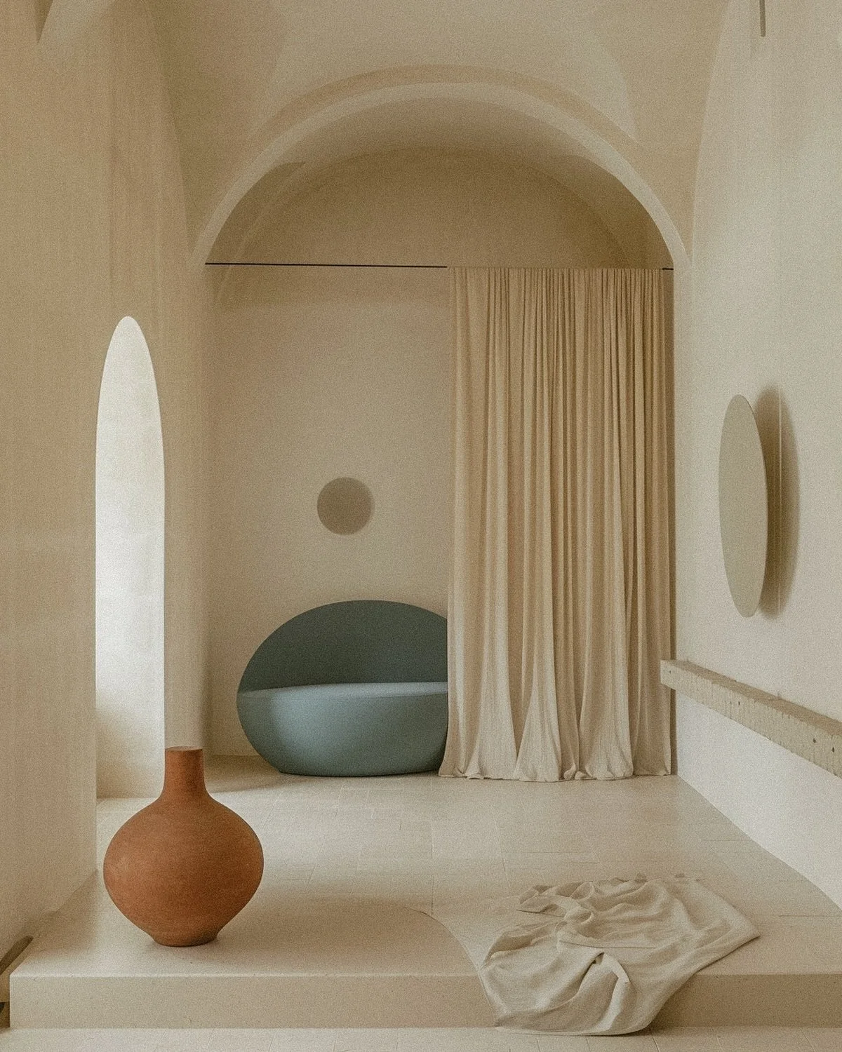 Minimalist interior with a tall, rounded arch window, beige curtain, geometric decor including a round wall hanging, large globe-shaped object, a ceramic vase, and a fabric piece on a ledge.