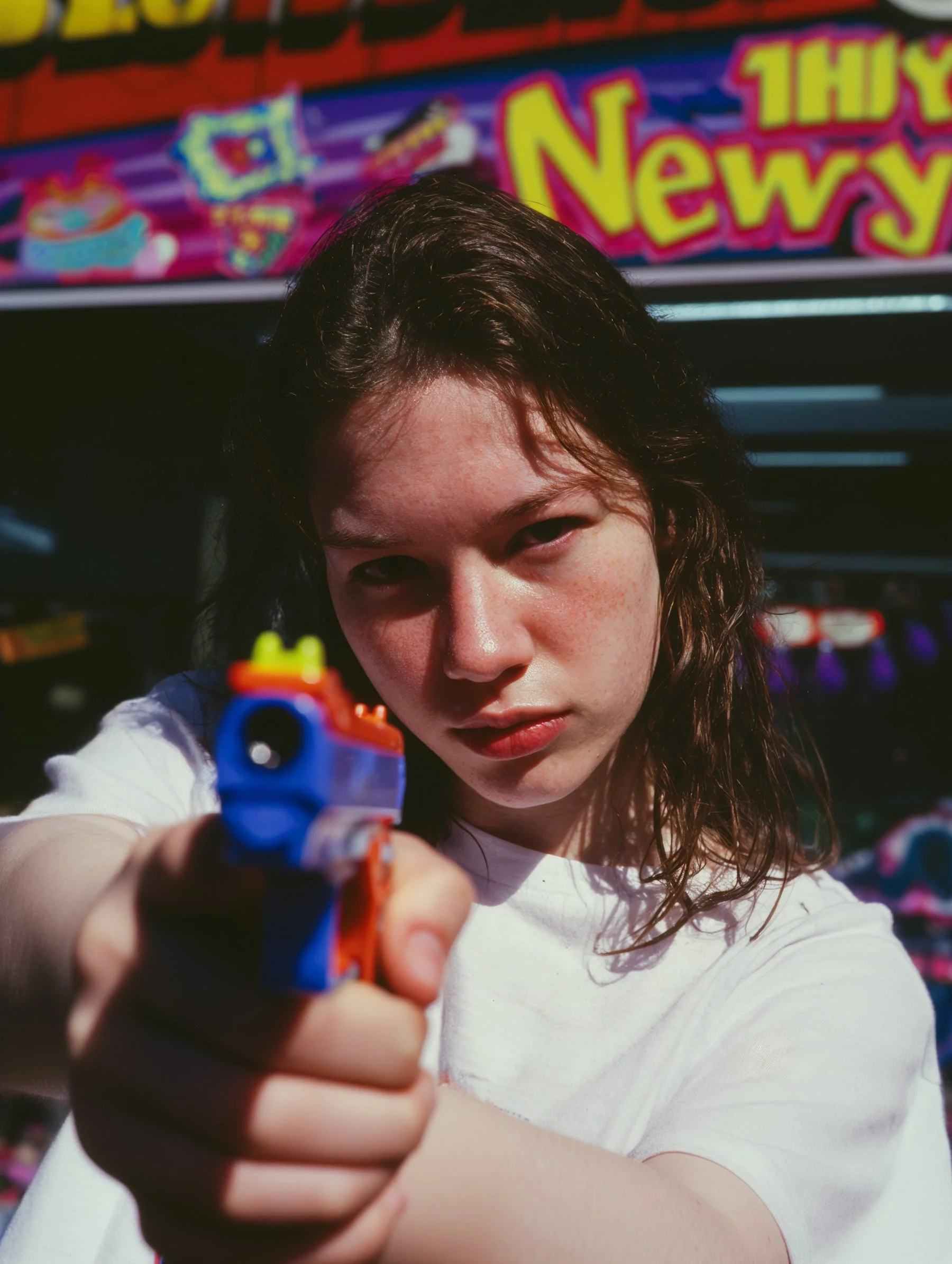 A young woman with wavy brown hair aiming a colorful toy gun directly at the camera, set against a background of a toy store with bright signs and shelves of toys.
