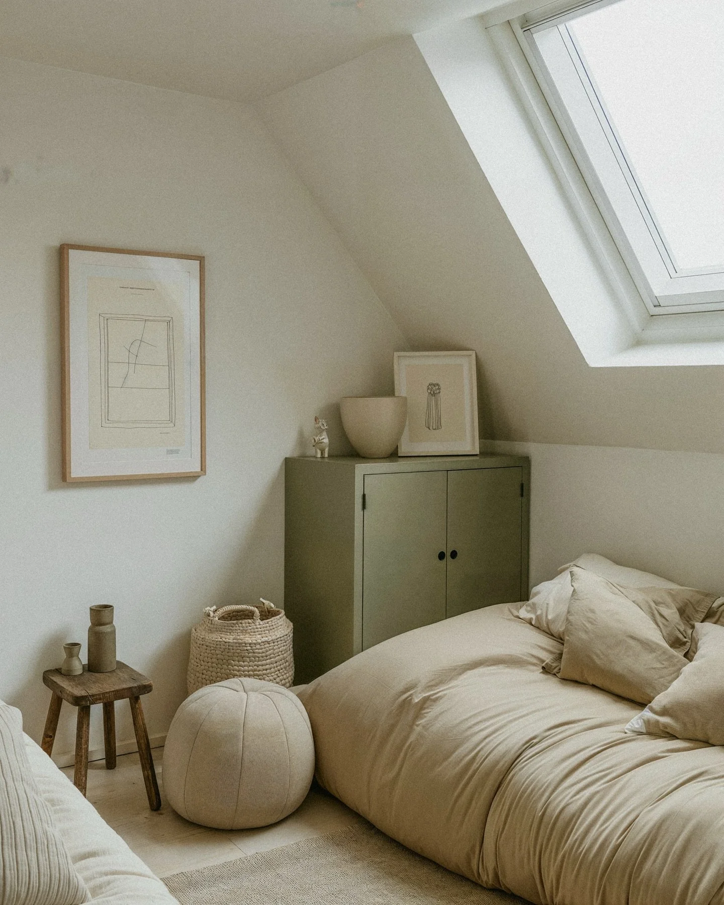 A cozy bedroom with a sloped ceiling, featuring a bed with beige bedding, a small green cabinet with decorative objects, a framed artwork and a skylight window.