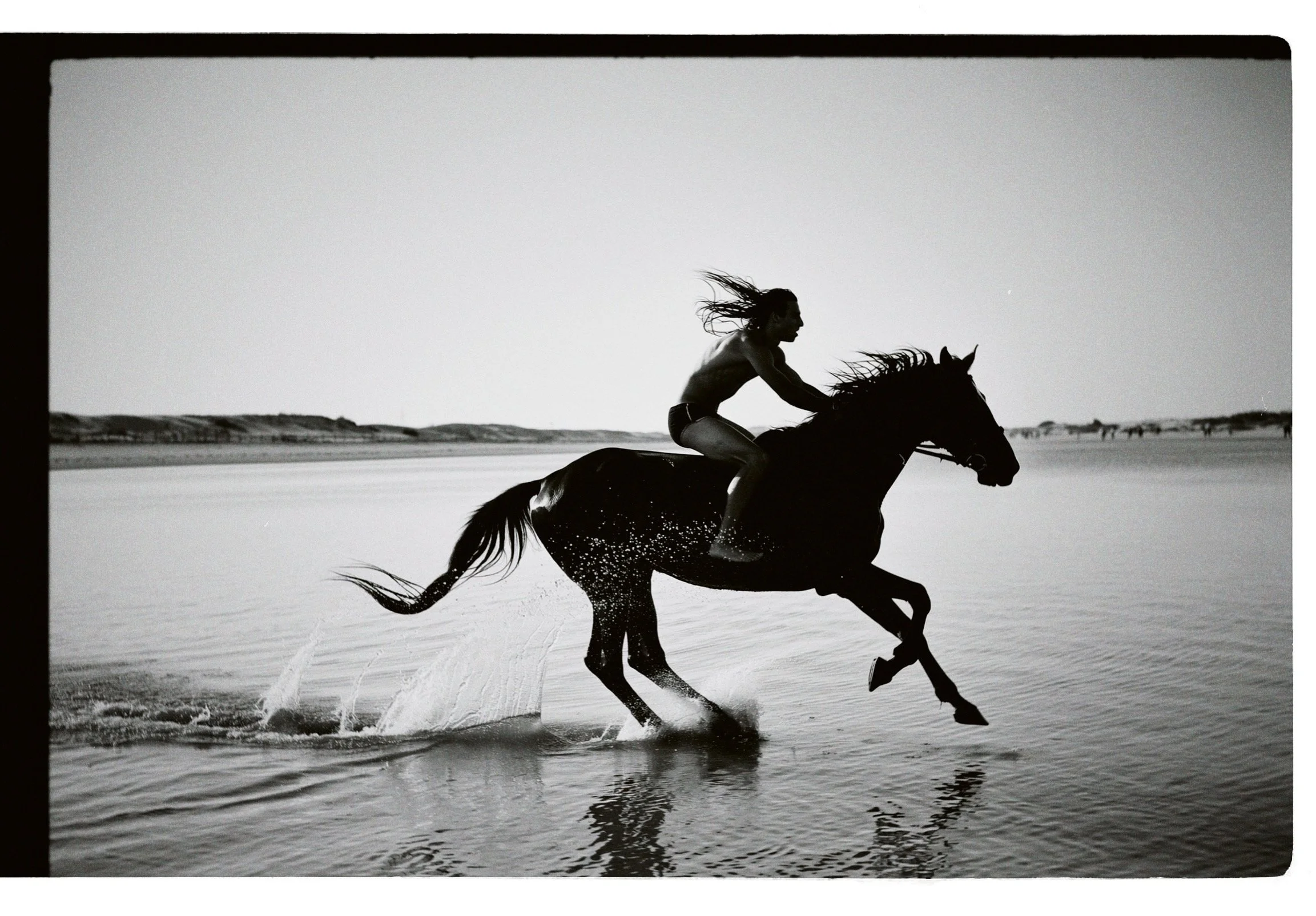 A person riding a horse through water on a beach, captured in black and white.
