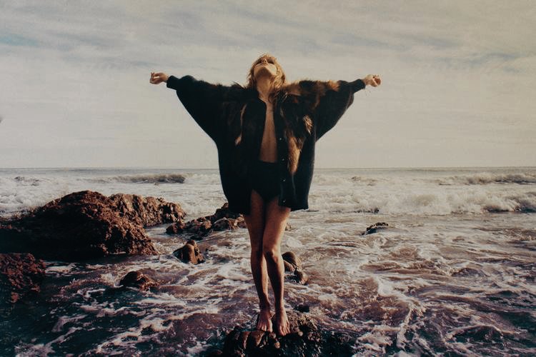 Woman standing on rocks at the beach with arms outstretched and eyes closed, wearing a fur coat with wet hair and ocean waves in the background.