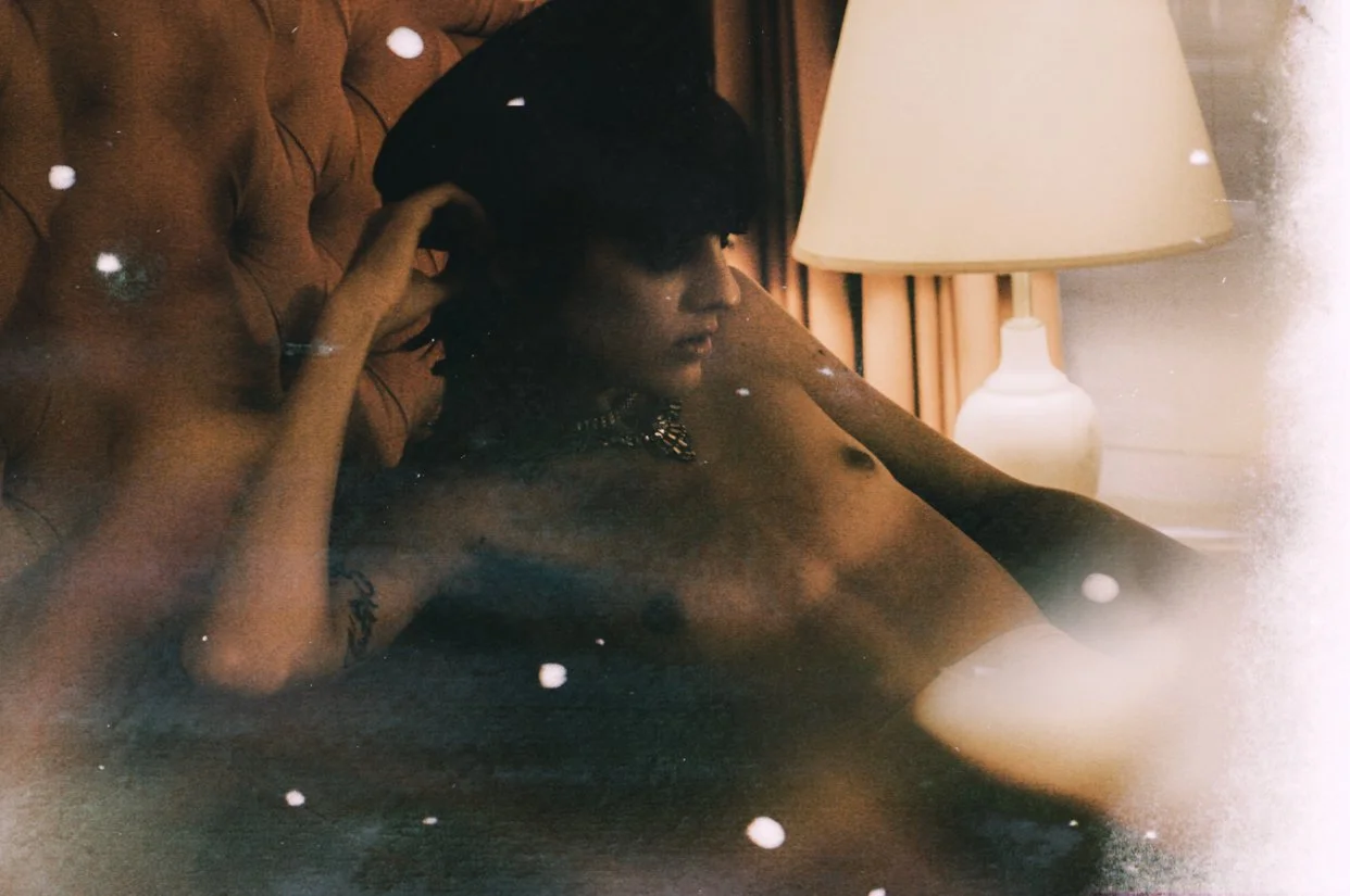 A man with tattoos lying on a brown tufted headboard, with a beige lamp on a side table in the background. He is shirtless, has dark hair, and is touching his hair with one hand, looking to the side. There are white specks and light effects added to 