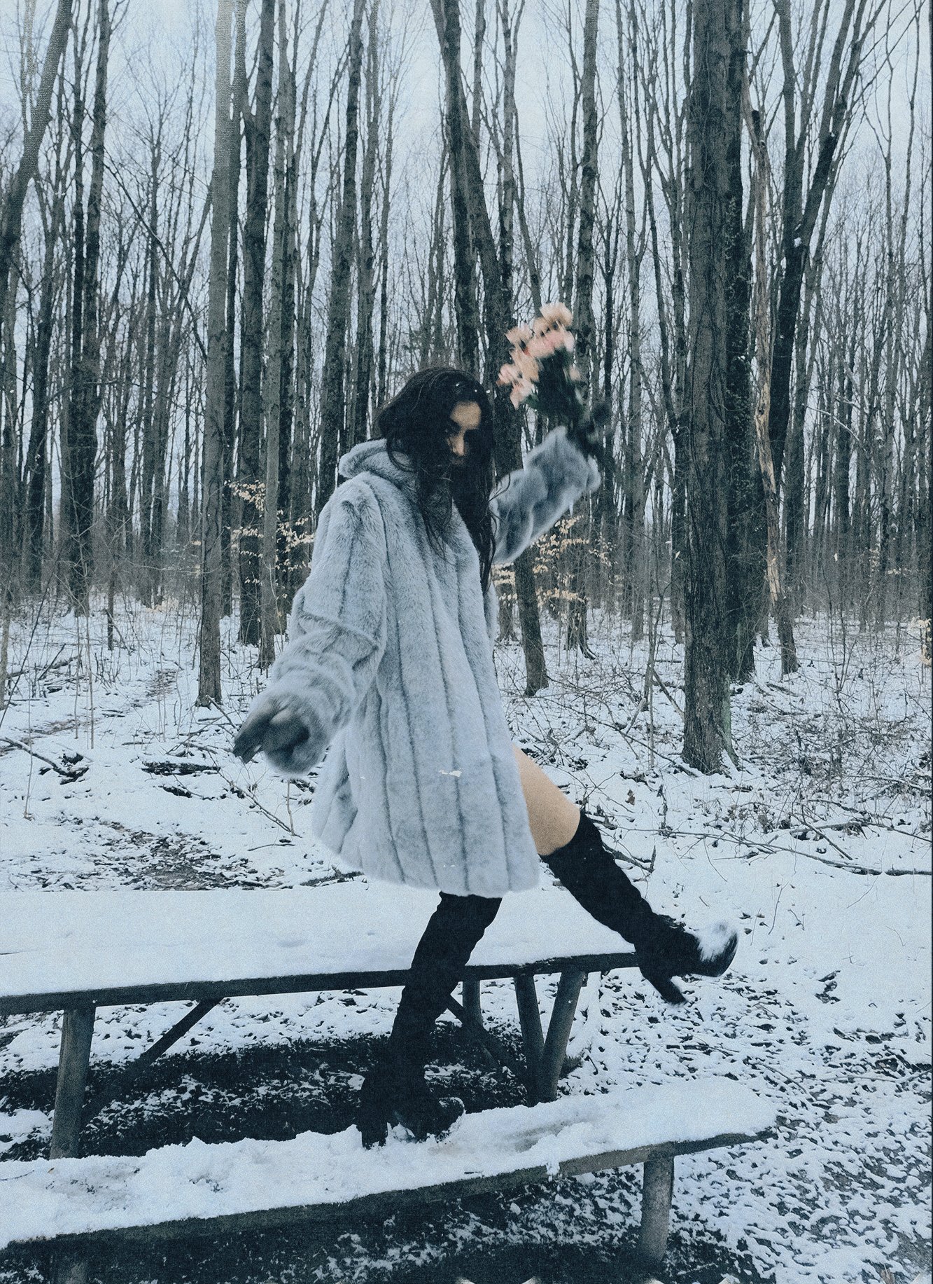 A woman in a long grey fur coat and black boots balancing on a snow-covered picnic table in a winter forest, holding flowers in one hand.