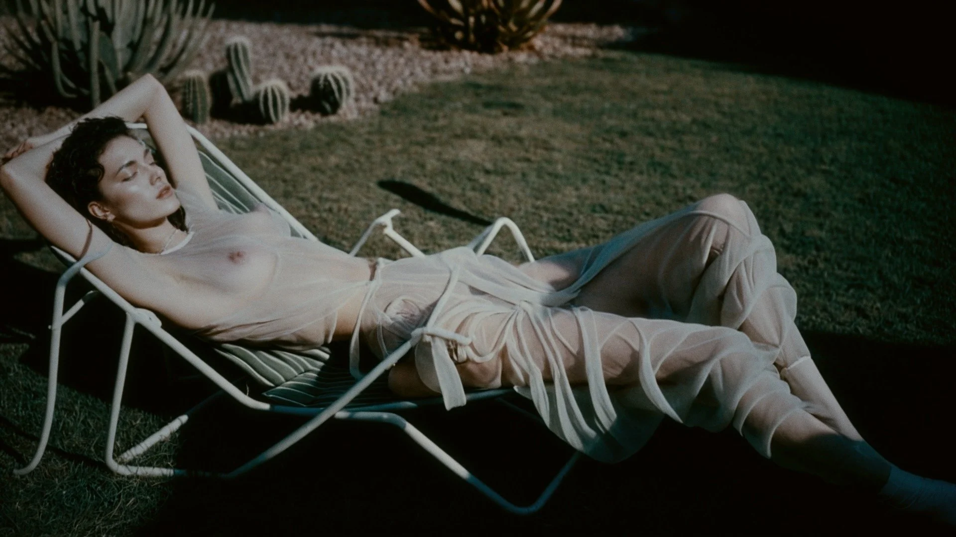 A woman with dark curly hair reclining on a striped lounge chair outdoors at night, wearing a sheer white dress, with cacti and desert plants in the background.