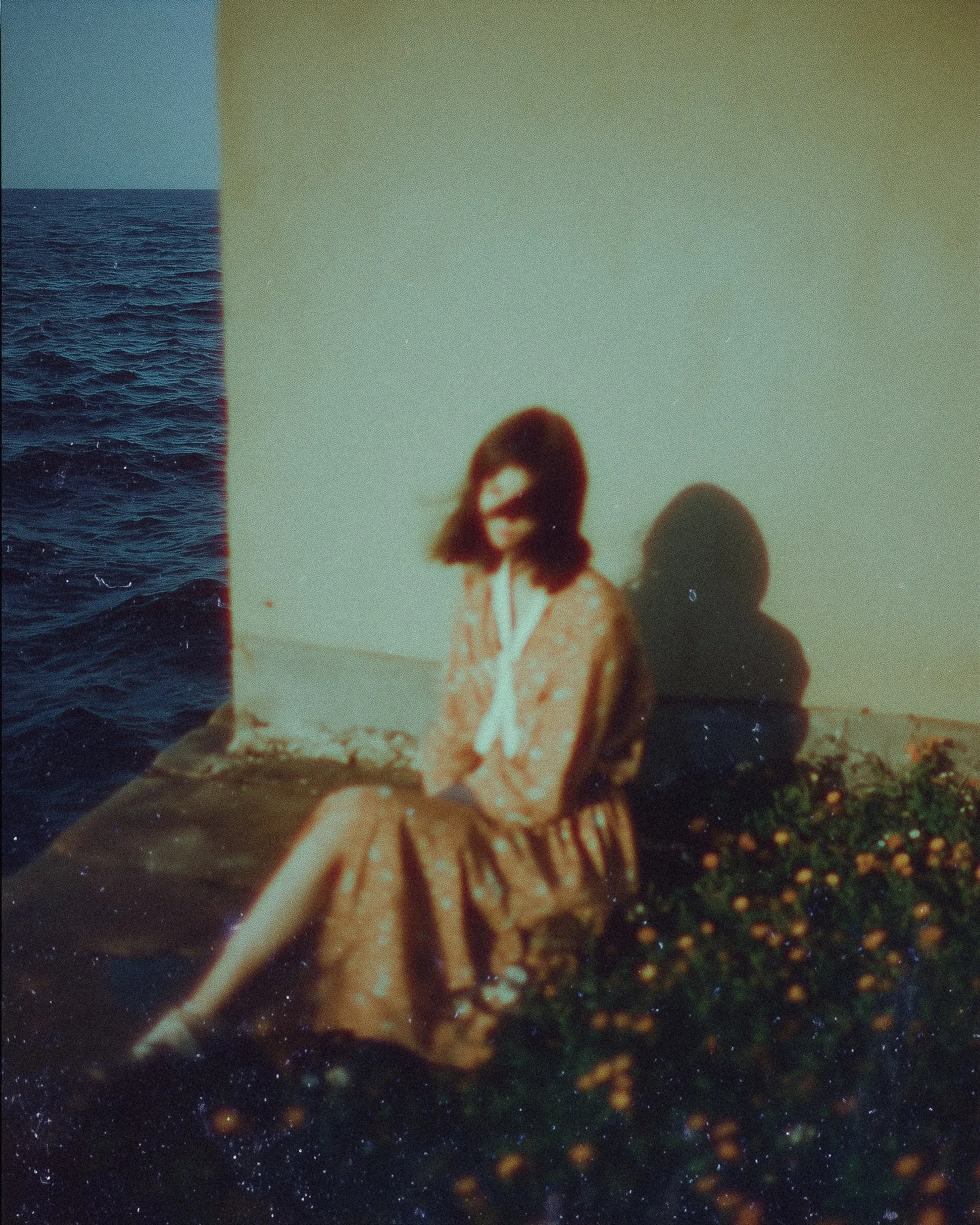 A woman wearing sunglasses and a patterned skirt sitting outdoors on a concrete surface next to a wall with a large shadow cast behind her. The scene appears vintage or analog-style with soft focus and warm tones.