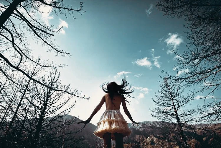 A girl in a tan dress jumps with her back to the camera in an outdoor setting with leafless trees and mountains in the background on a partly cloudy sky.