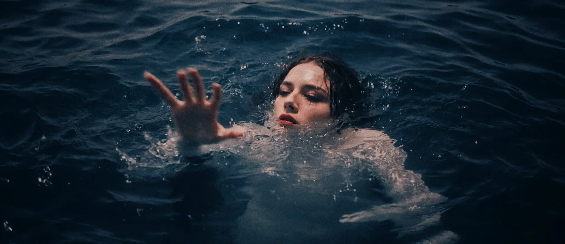 Woman with dark hair swimming in dark blue water, reaching out with her hand.