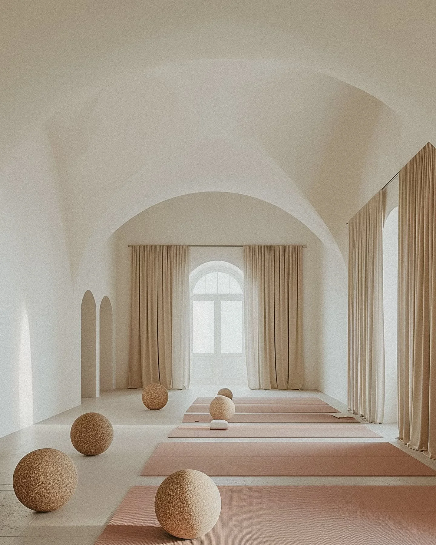 Empty yoga studio with pink mats, large beige textured balls, and cream curtains in front of arched windows.