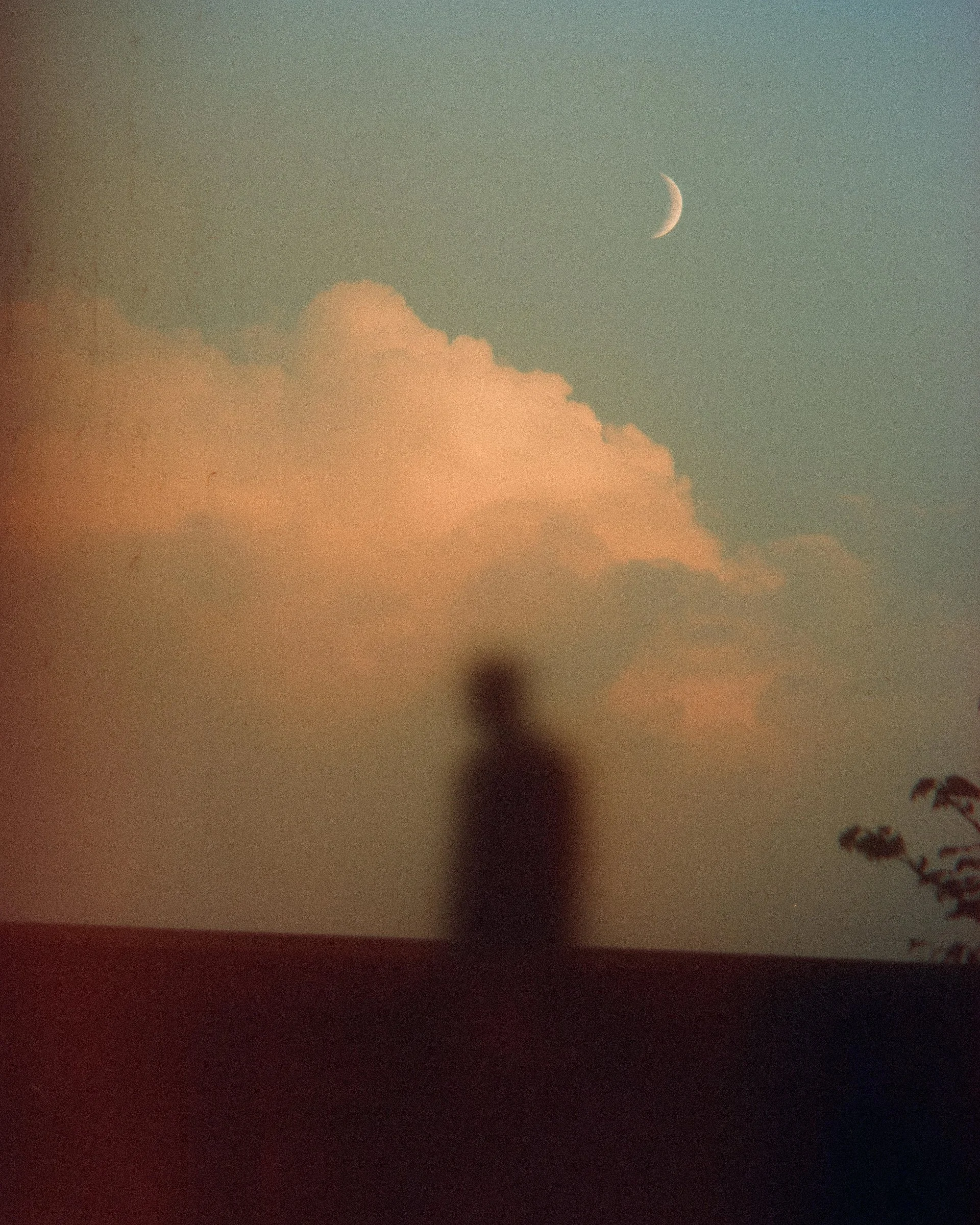 Blurred silhouette of a person standing outdoors at sunset or sunrise, with a large cloud and a crescent moon in the sky.