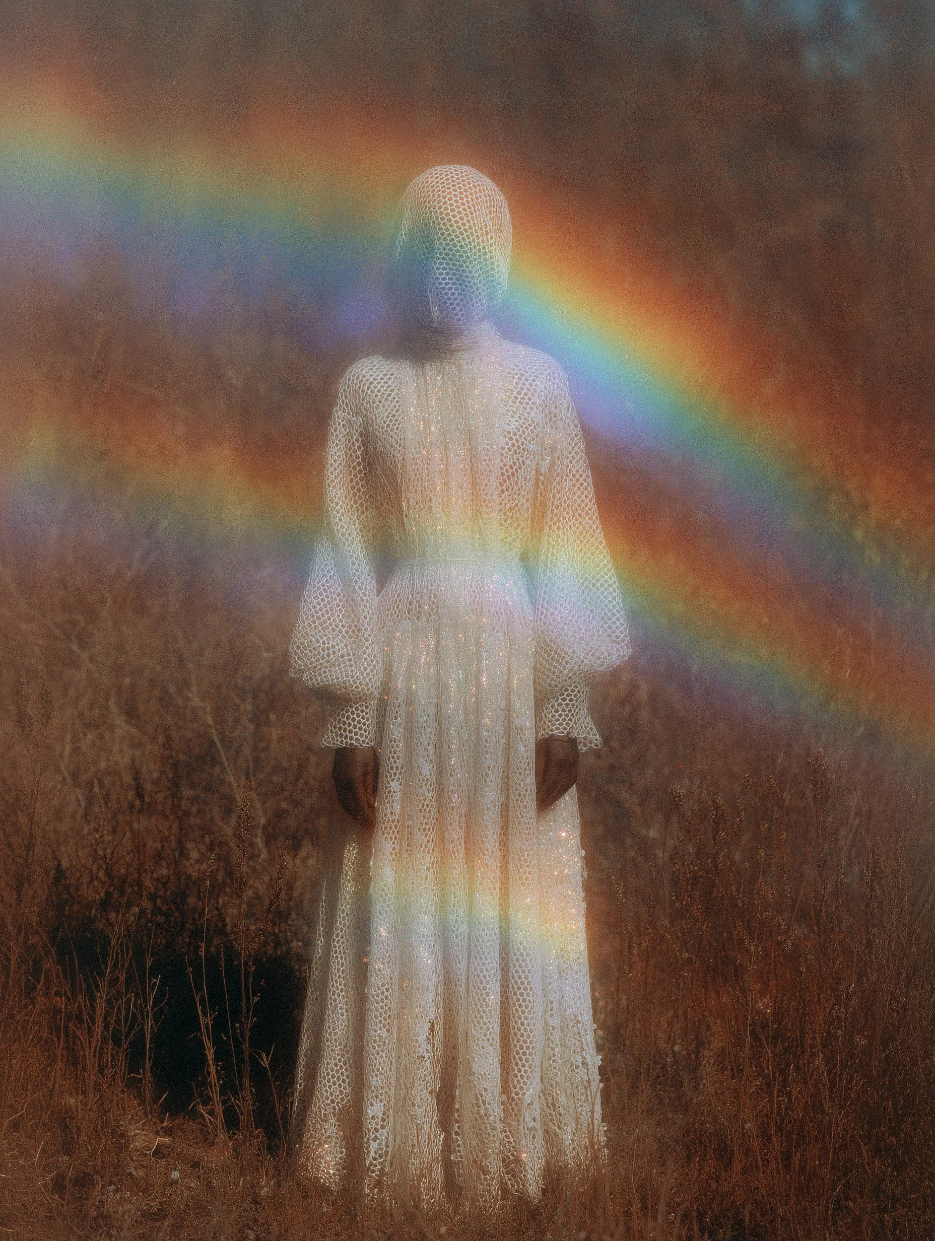 A person in a white, long, shimmering dress stands in a field with dry grass, surrounded by a rainbow and a cloudy sky.
