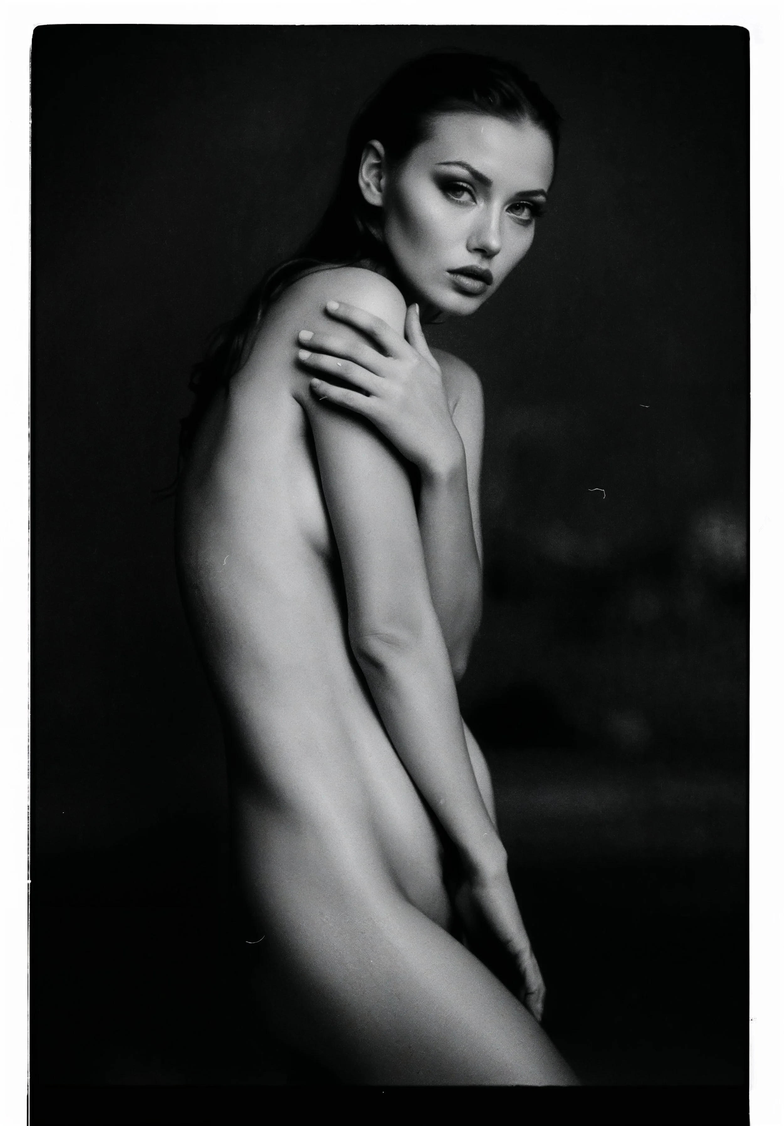 Black and white photograph of a young woman with long hair, topless, covering her chest with her arms, looking directly at the camera with an intense expression.