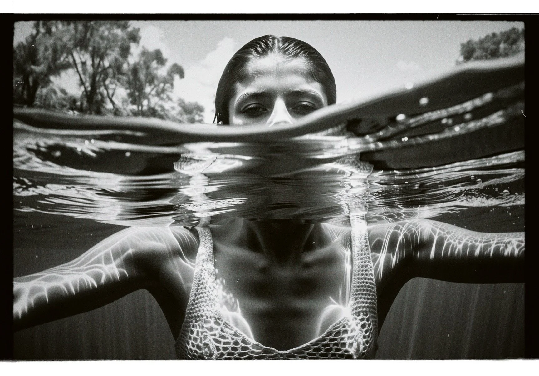 Black and white photograph of a woman partially submerged in water, with only her face and shoulders visible above the water surface. The woman is wearing a textured top, and the sunlight creates reflections on her skin. The background shows trees an