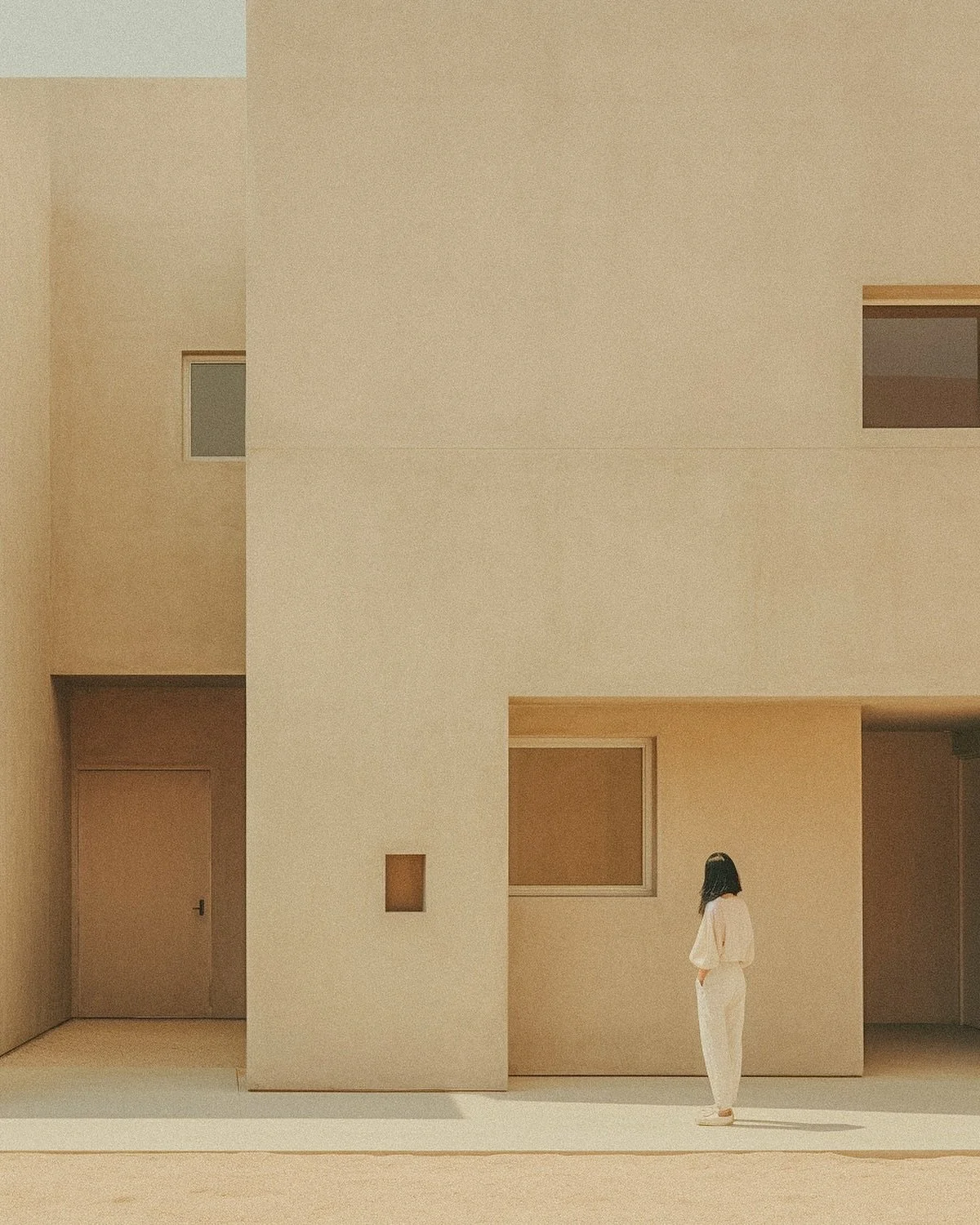 A woman with long dark hair, dressed in white, stands in front of a modern beige building with minimalistic architecture, including rectangular windows and doors, on a sunny day.