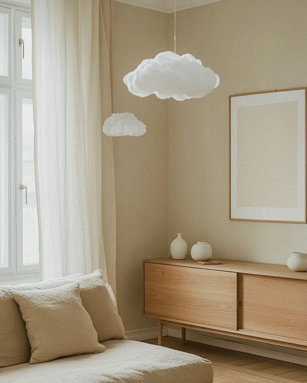 A cozy, minimalistic living room with beige walls, sheer white curtains, a beige couch with pillows, a wooden sideboard with white vases, a large blank picture frame, and calendar-shaped cloud pendant lights hanging from the ceiling.