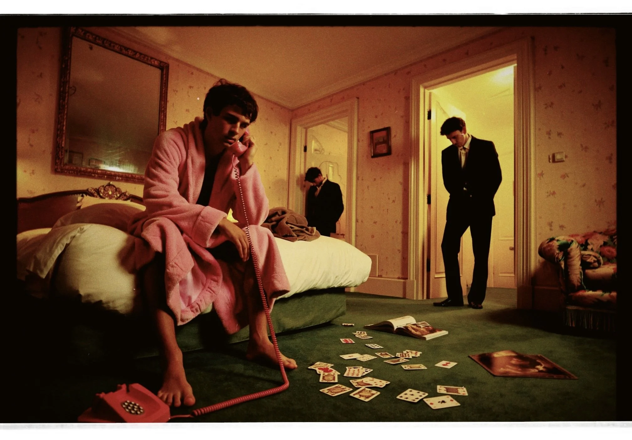 A young man in a pink bathrobe sitting on a bed, talking on a red rotary phone, with scattered playing cards and open magazines on the green carpeted floor. Two other men in suits are standing in the doorway and in the doorway of an adjacent room in 
