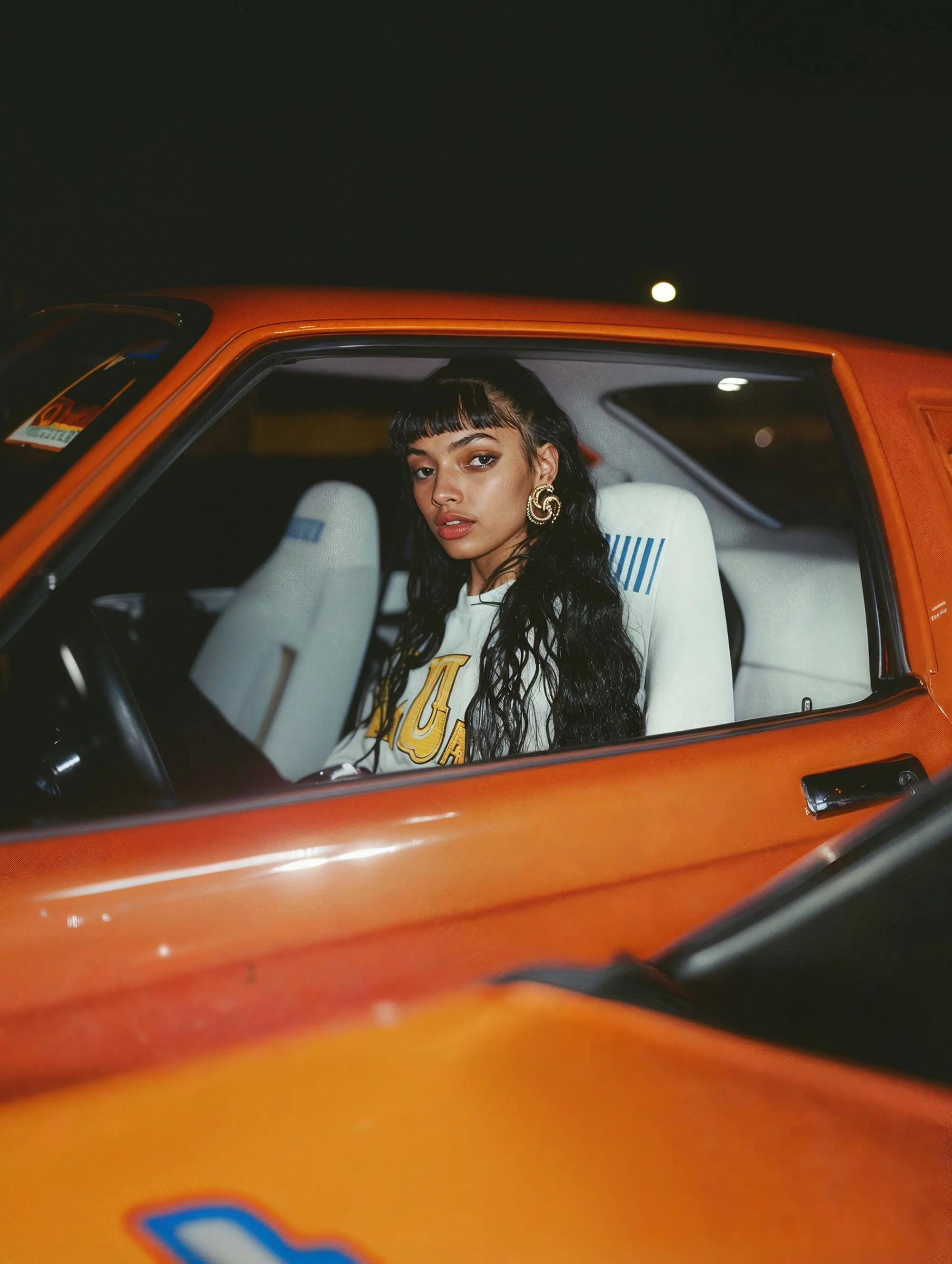 A woman with long black curly hair and gold earrings sitting in an orange vintage car at night, looking at the camera.