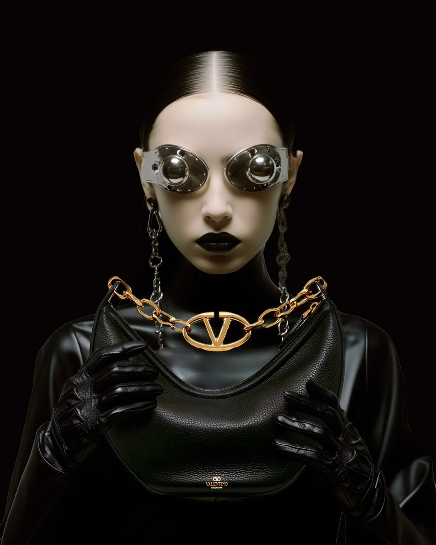 Fashion model with chrome goggles, black lipstick, black gloves, Valentino handbag, and chunky gold chain necklace with Valentino logo, against a black background.