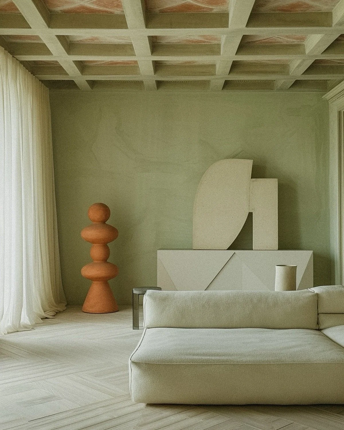 Minimalist living room with cream-colored sofa, abstract white sculptures on a sideboard, a tall terracotta sculpture resembling stacked geometric shapes, and light-colored curtains.