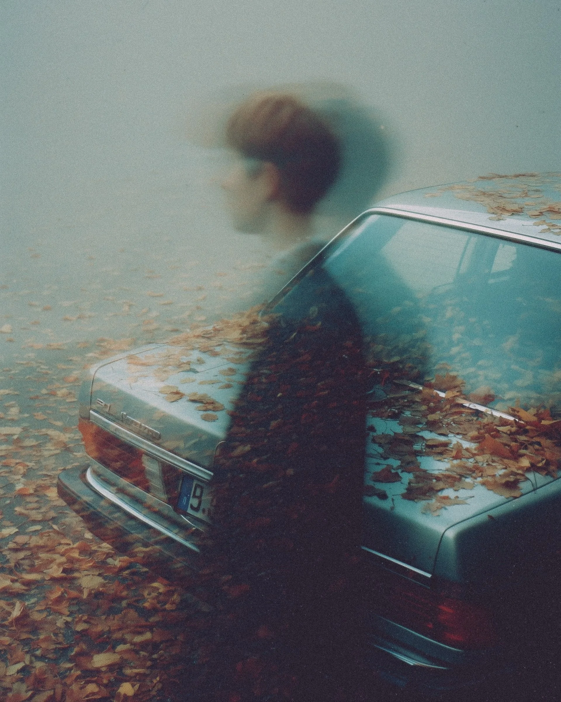 A double exposure photograph showing a partial view of a person's upper body and head, with a reflection of an autumn scene on the back of a car, which is also covered with fallen leaves.
