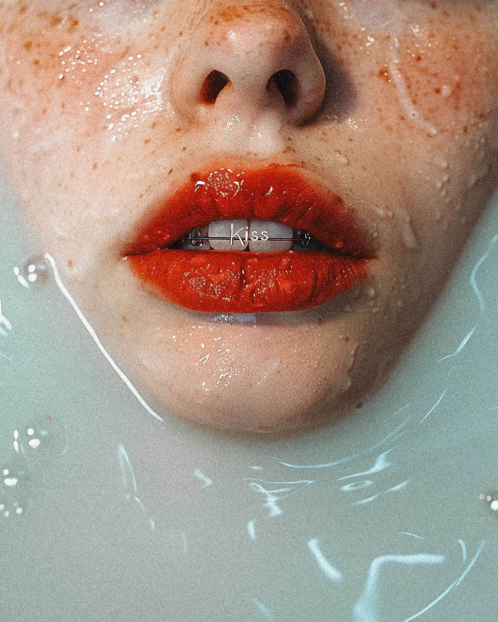 Close-up of a person's lips with red lipstick, teeth with a 'Kiss' dental grill, water droplets on face, and water surrounding the face.
