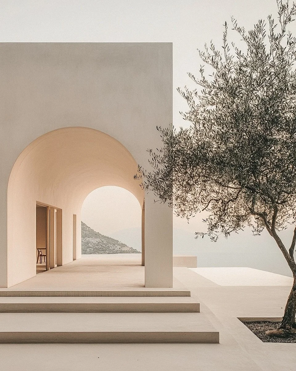 Minimalist modern building with arched entryway, steps leading up, and a tree to the right, overlooking a distant hill and ocean.