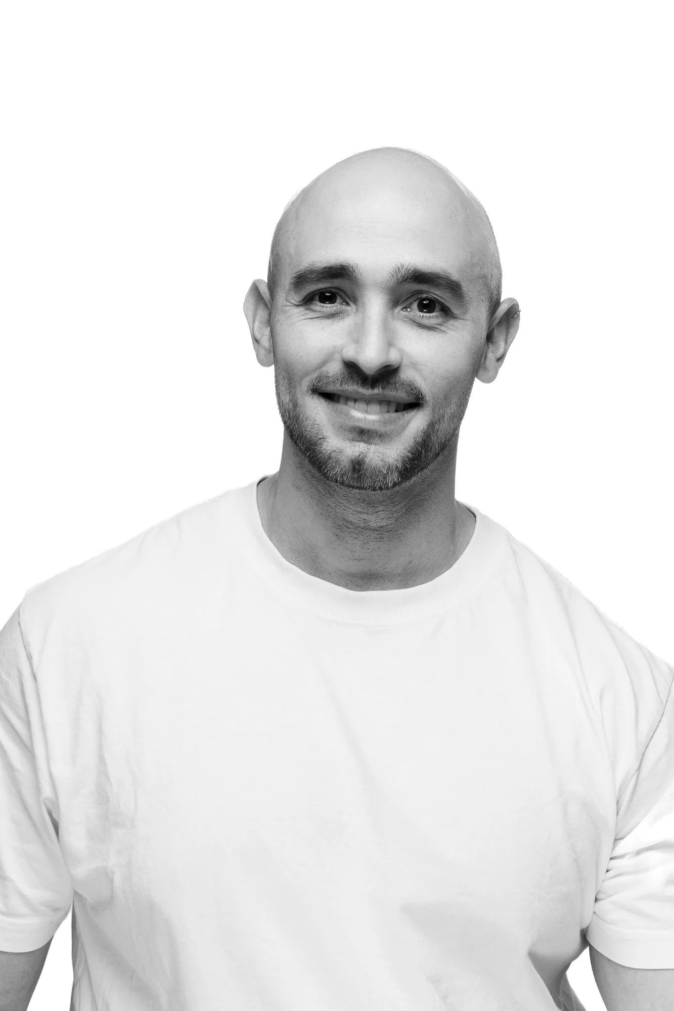 Black and white portrait of a smiling man with a beard and shaved head, wearing a plain white t-shirt, against a plain white background.