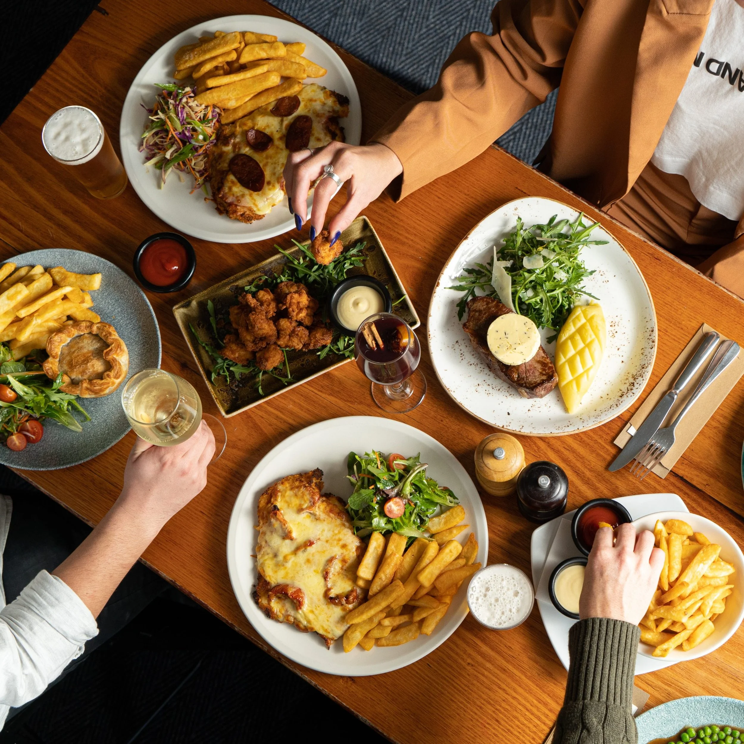A table set with various plates of food including salads, French fries, a bread bowl with soup, grilled meat with mashed potatoes, fried chicken with dipping sauces, and beverages like beer and wine