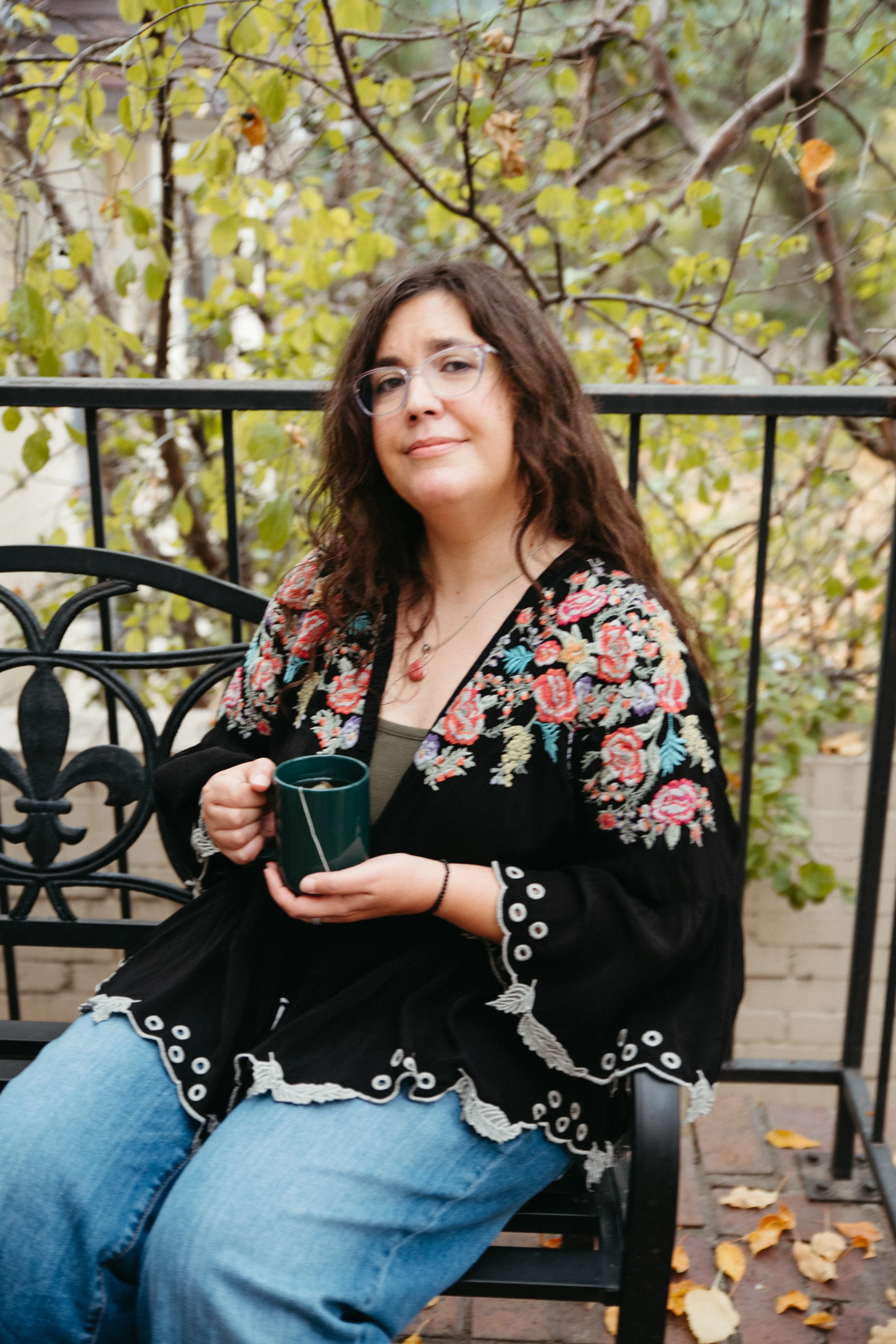 A woman with glasses, wavy brown hair, and a floral embroidered black jacket sitting on a black iron chair outdoors, holding a teal mug, with fallen autumn leaves on the ground and leafy trees in the background.