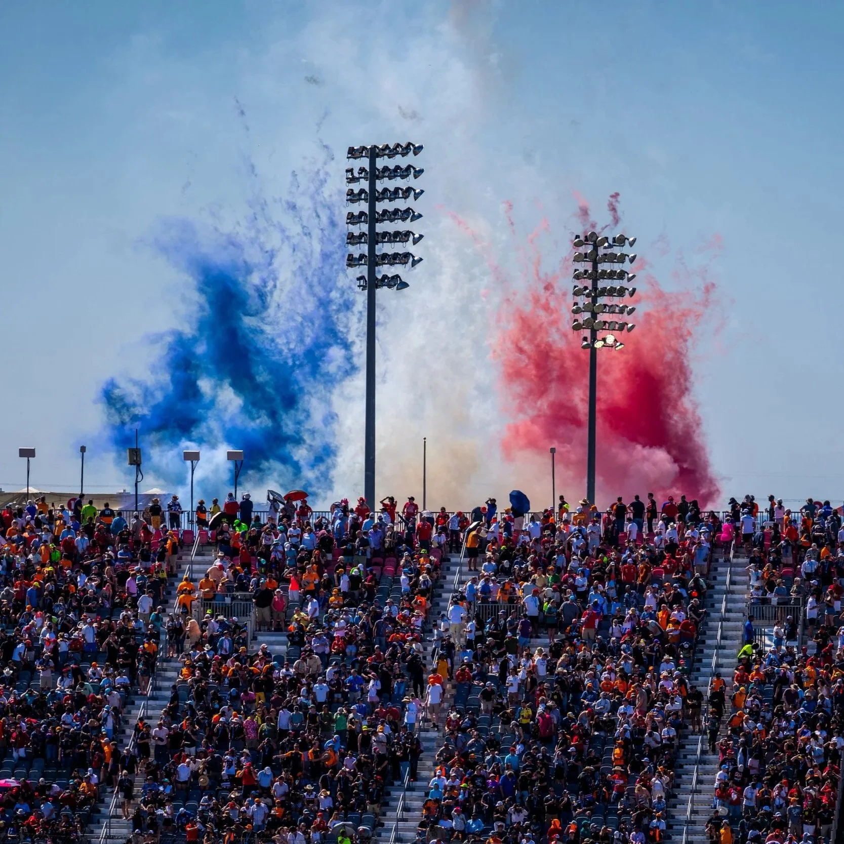 Crowd of people at a race track with red, white, and blue smoke in the background from fireworks or pyrotechnics.