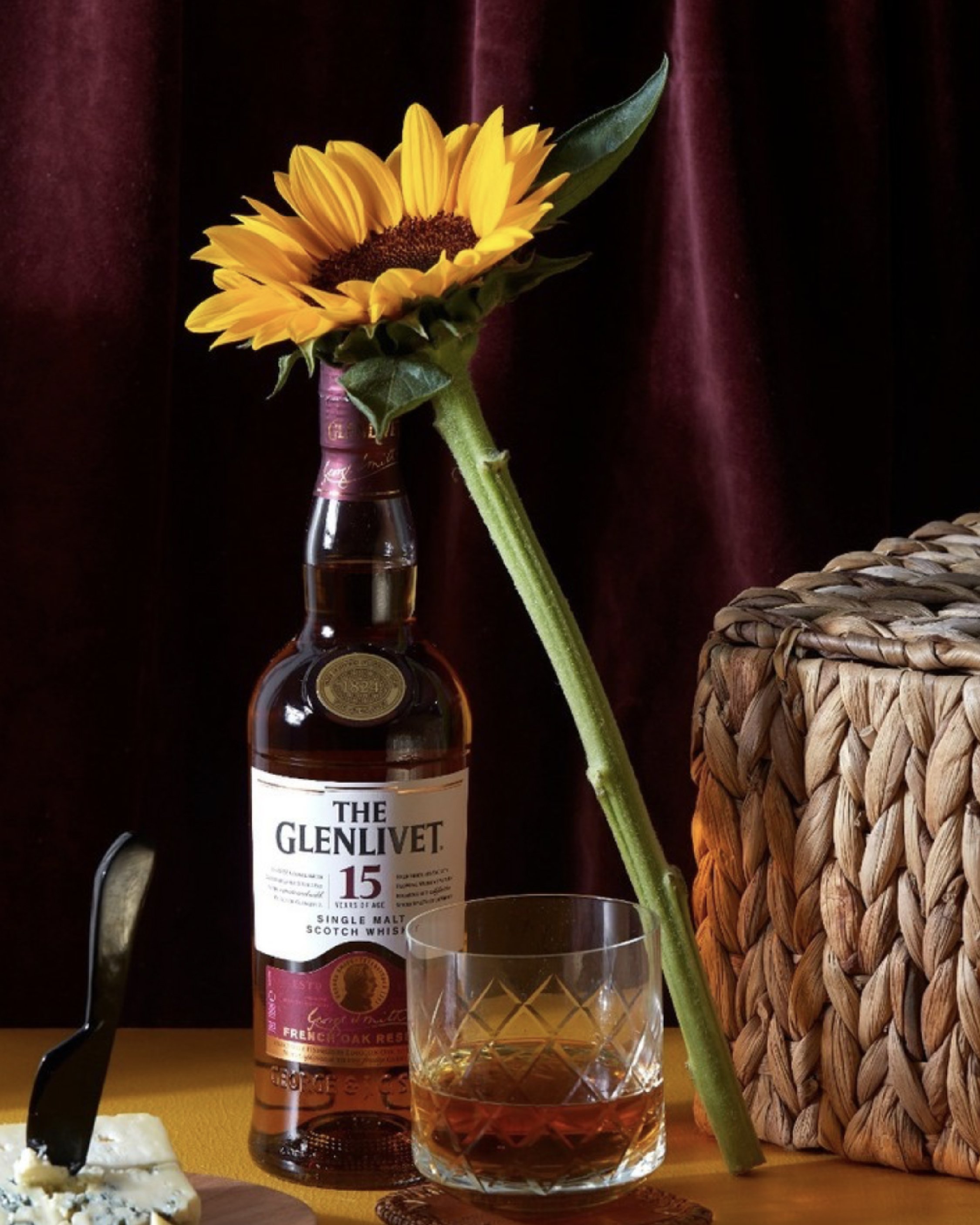 Bottle of Glenlivet 15-year-old whisky with sunflower, glass of whisky, wicker basket, and cheese on a dark red background.