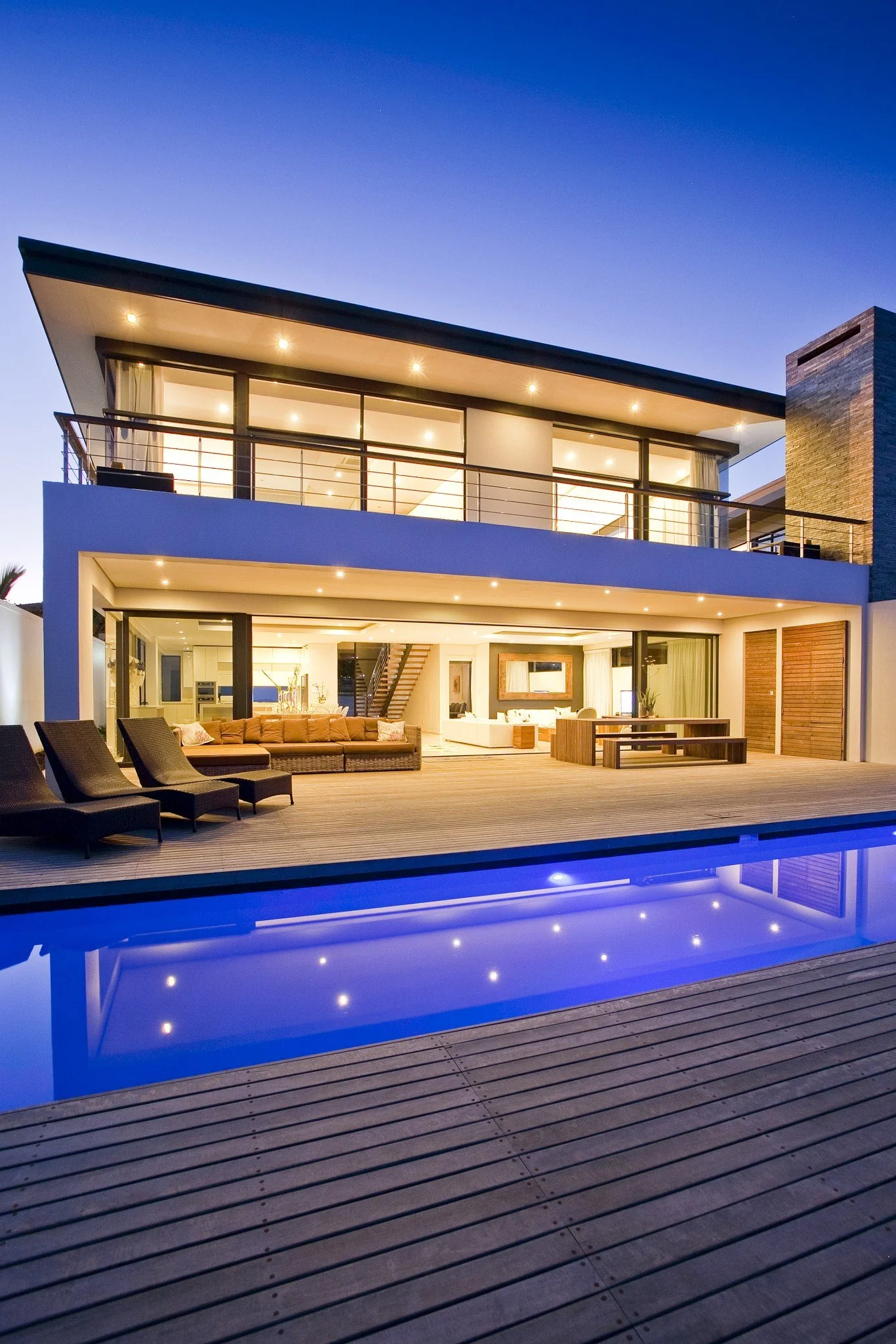 A timber deck is lit up by the setting sun , there are dining tables and loungers on the timber deck which surrounds a pool and leads to the lounge