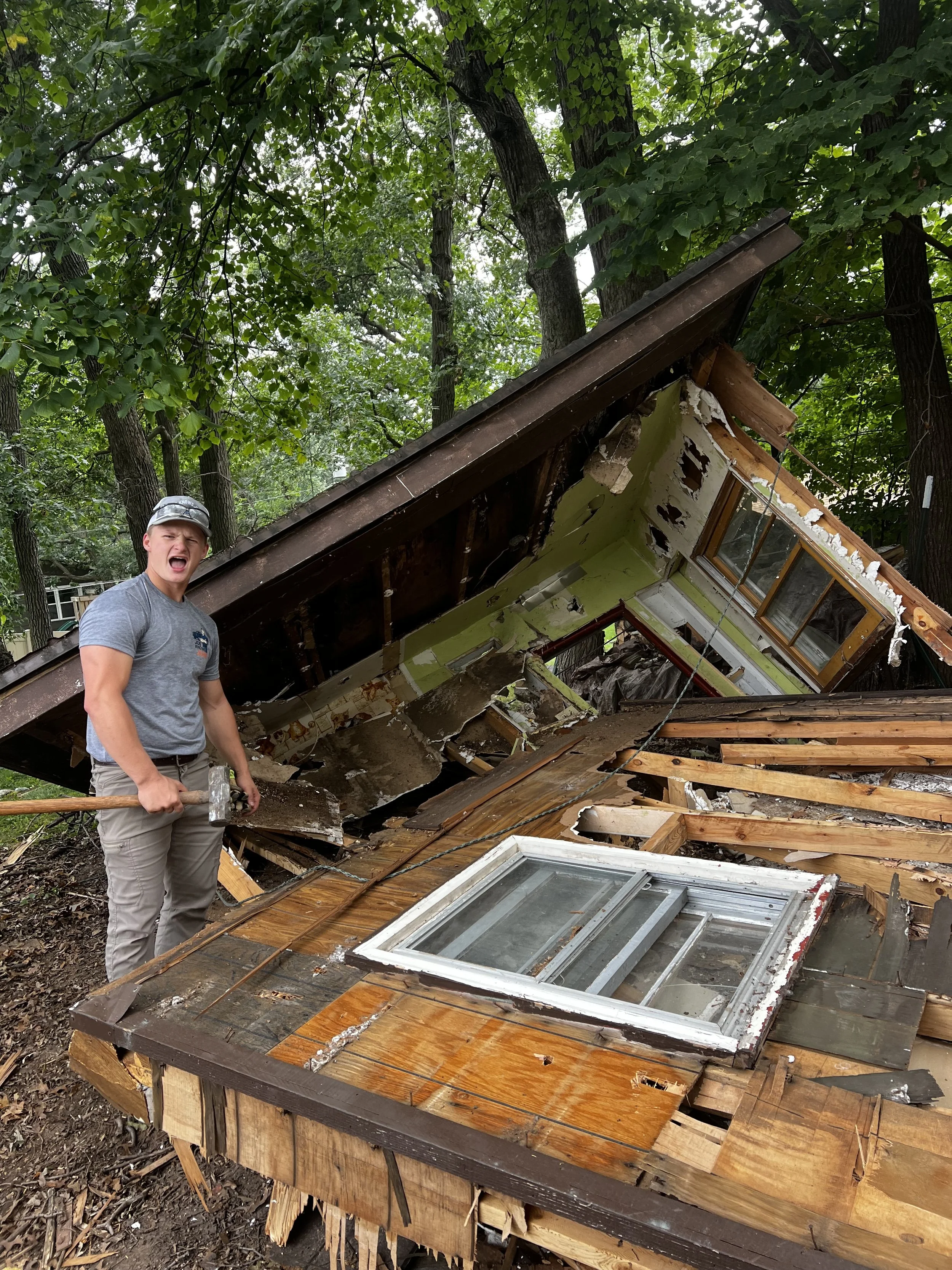 A man standing next to a heavily damaged house, holding a hammer, with debris and broken wooden planks, and a fallen roof in a wooded area.