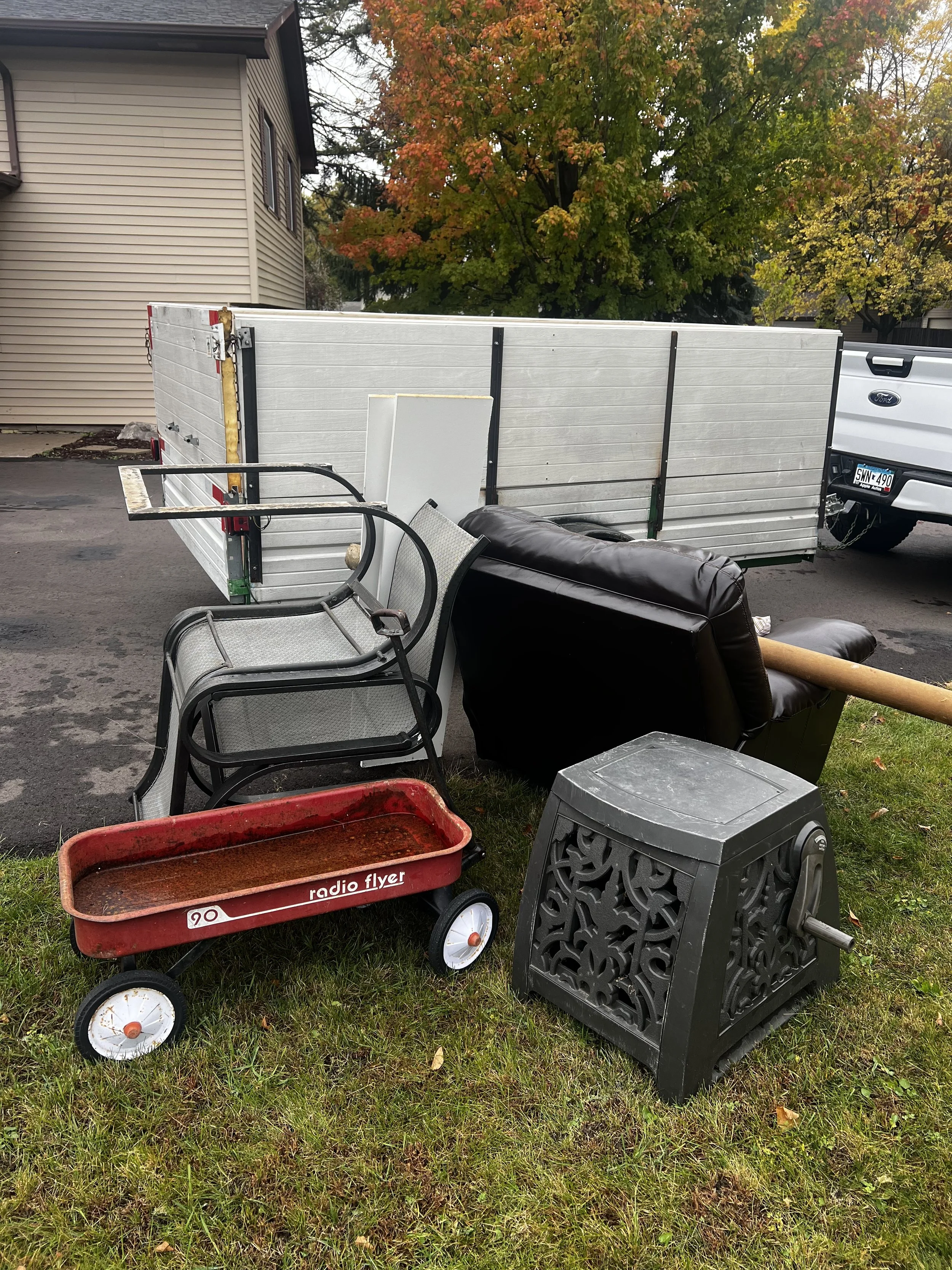 Various pieces of outdoor furniture and items, including a red Radio Flyer wagon, black chairs, a black patio table, a black stool, a white panel, a black decorative cube, a brown leather couch, and a white fence, placed on a grassy lawn with a house and trees in the background.